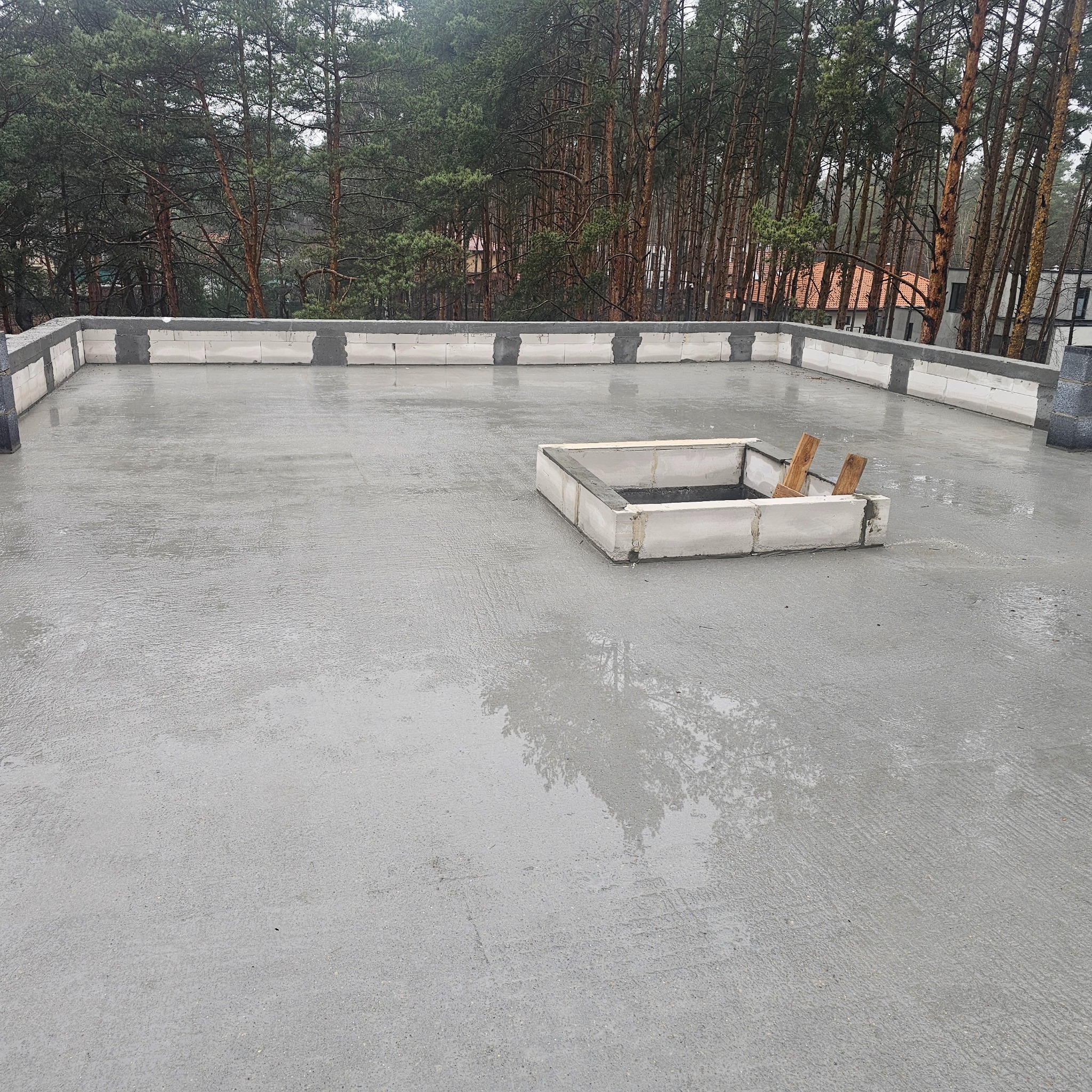 Świeżo wylana, mokra betonowa płyta dachu z obrzeżem z bloczków betonowych i małym kwadratowym otworem w płycie, na tle las sosnowy.