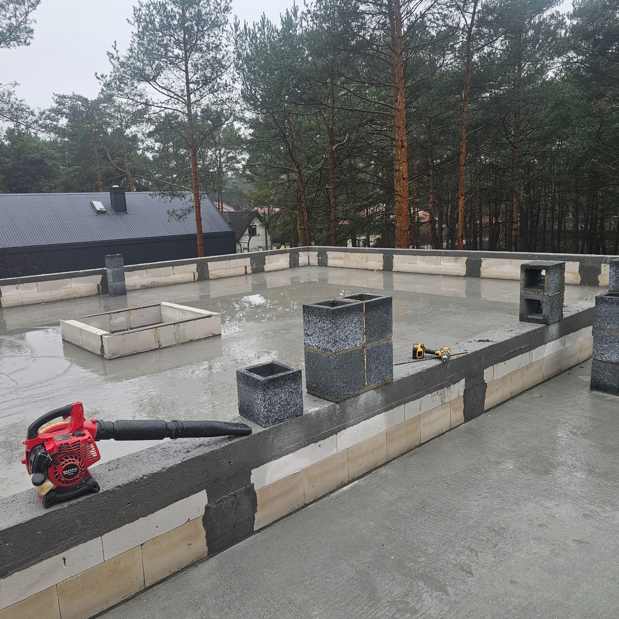 Budowa dachu płaskiego: wylany beton, bloczki betonowe, kominy wentylacyjne w trakcie wznoszenia, dmuchawa do liści Bavaria i wkrętarka akumulatorowa DeWalt na mokrej powierzchni.