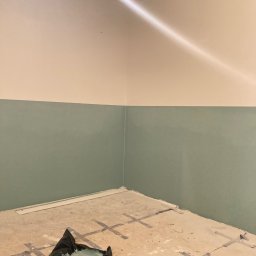 Rafał Potok - Świeżo pomalowane ściany w dwóch odcieniach: kremowym u góry i pastelowym zielonym na dole, widoczne ślady taśmy malarskiej na podłodze, puszka farby i zabrudzona folia malarska w rogu pomieszczenia.