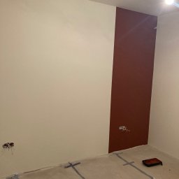 Rafał Potok - Wnętrze pokoju w trakcie malowania: ściany częściowo pokryte beżową i bordową farbą, zabezpieczona podłoga taśmą malarską, widoczna puszka elektryczna z wystającymi przewodami i wałek do malowania...