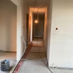 Korytarz w trakcie wykańczania, widoczne ściany z płyt kartonowo-gipsowych, instalacje elektryczne prowadzone po podłodze, surowy beton i wisząca żarówka.