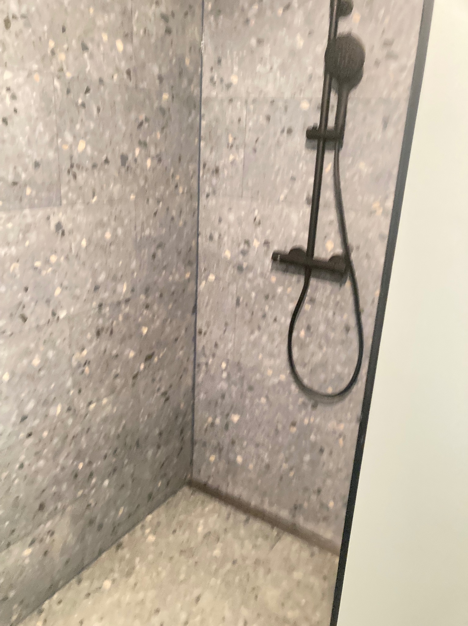 Wykończona kabina prysznicowa z szarymi płytkami lastryko na ścianach i podłodze, wyposażona w czarną armaturę natryskową.