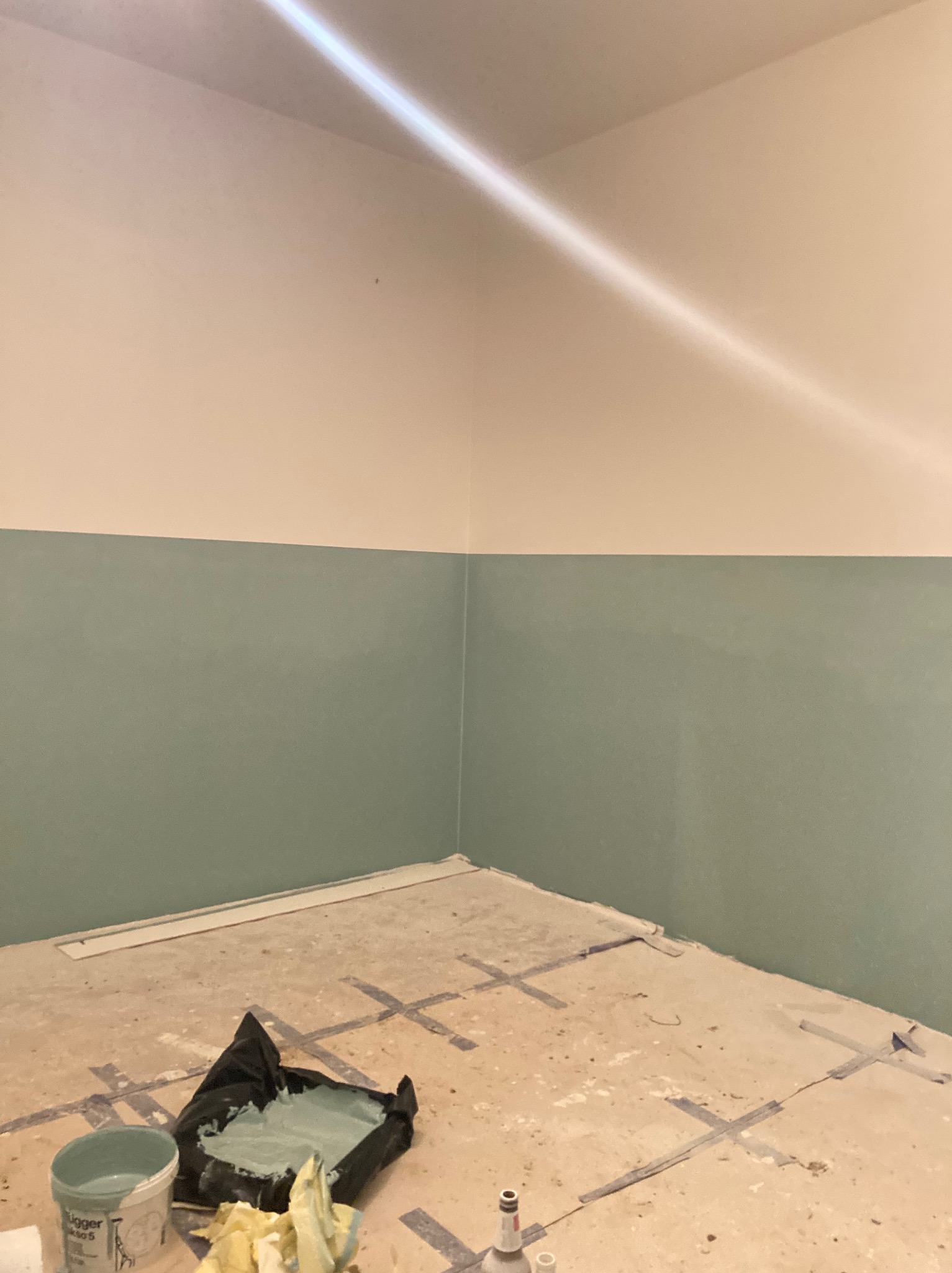Świeżo pomalowane ściany w dwóch odcieniach: kremowym u góry i pastelowym zielonym na dole, widoczne ślady taśmy malarskiej na podłodze, puszka farby i zabrudzona folia malarska w rogu pomieszczenia.