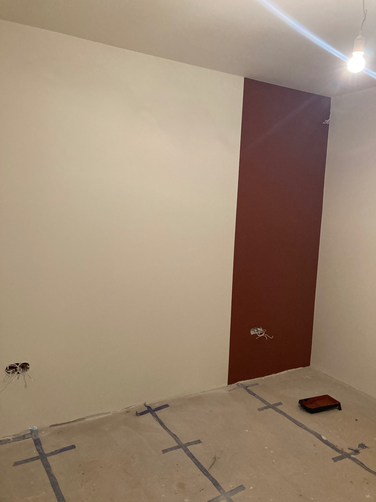 Wnętrze pokoju w trakcie malowania: ściany częściowo pokryte beżową i bordową farbą, zabezpieczona podłoga taśmą malarską, widoczna puszka elektryczna z wystającymi przewodami i wałek do malowania...