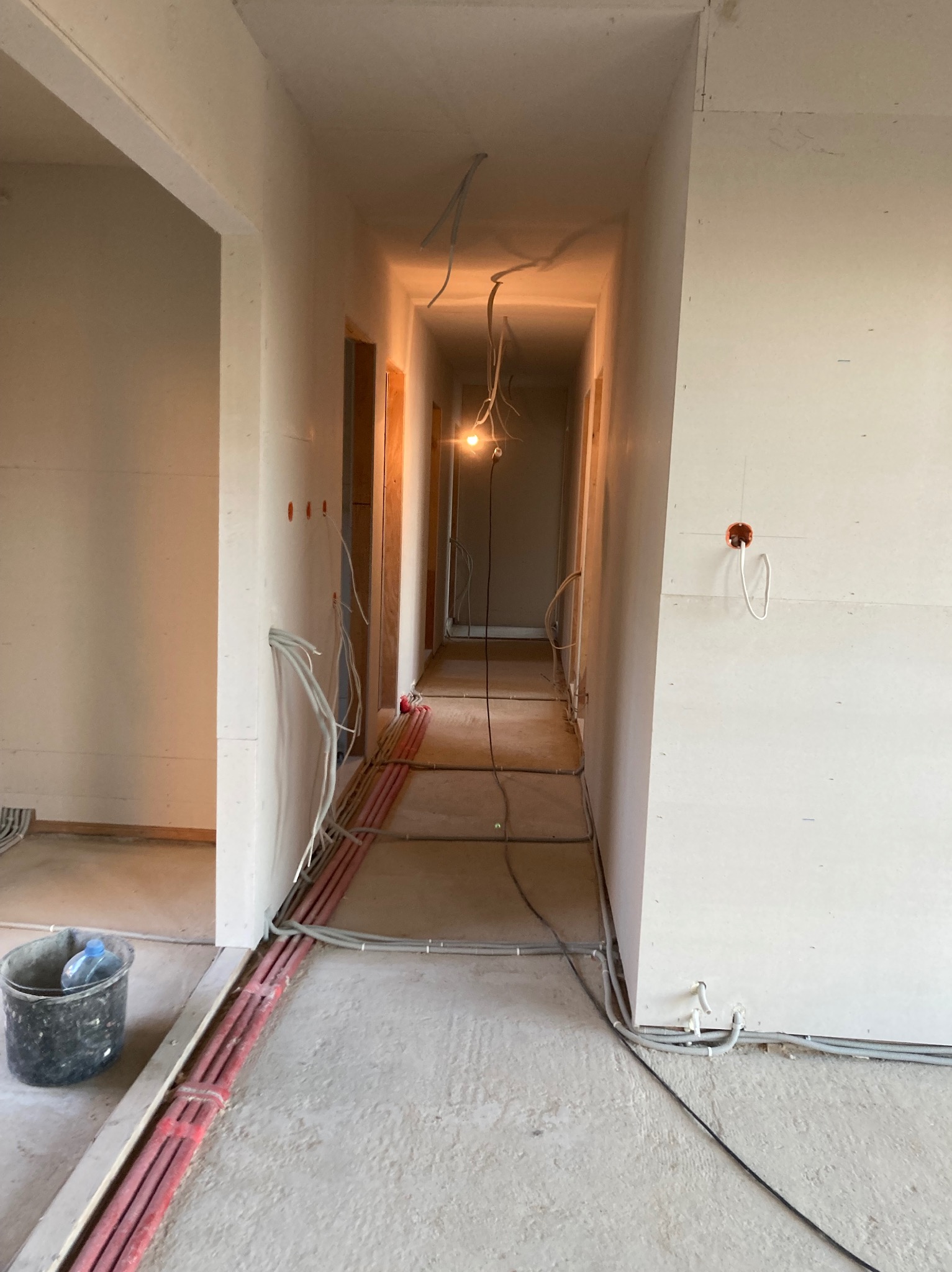 Korytarz w trakcie wykańczania, widoczne ściany z płyt kartonowo-gipsowych, instalacje elektryczne prowadzone po podłodze, surowy beton i wisząca żarówka.