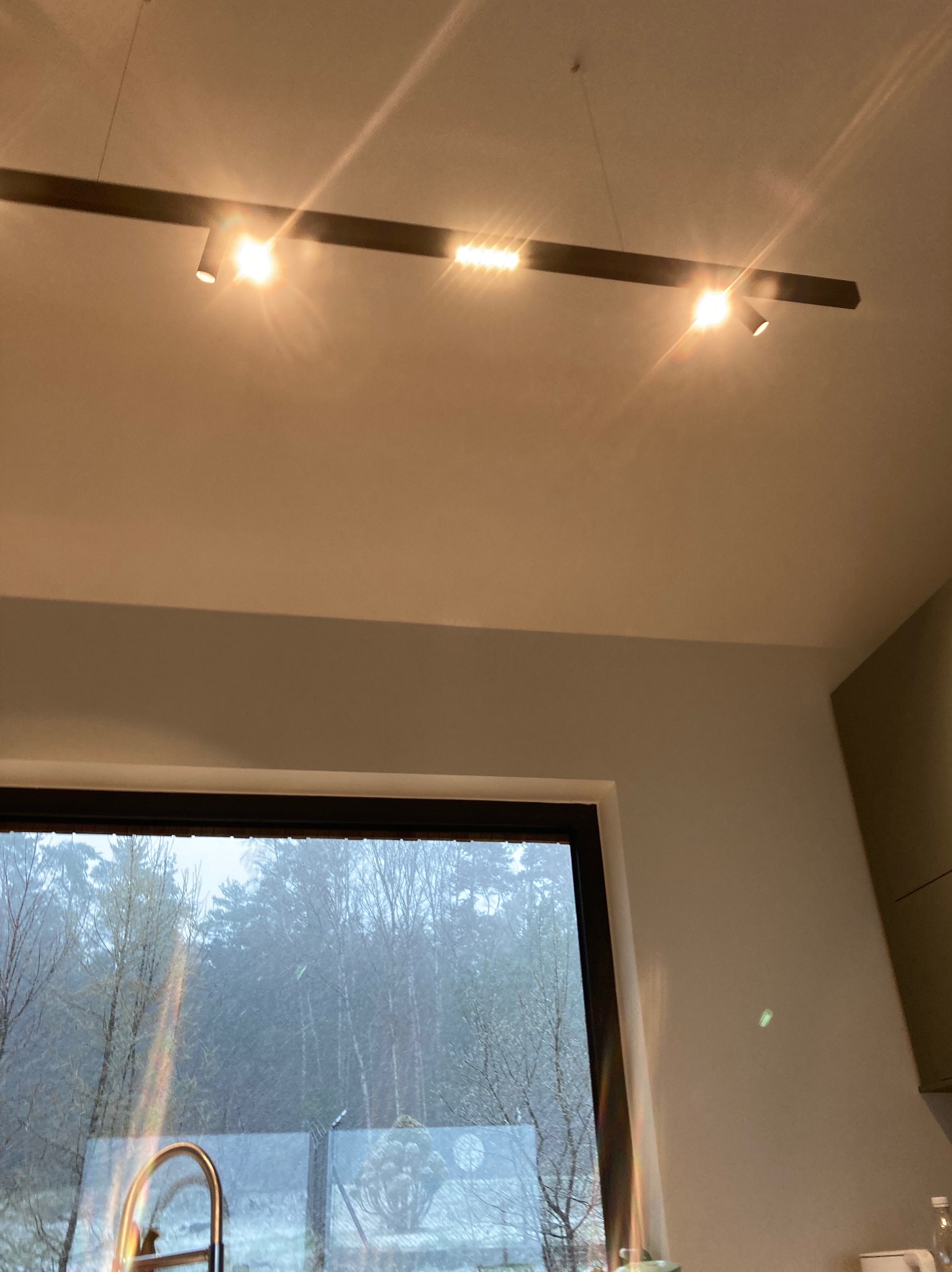 Nowoczesna listwa oświetleniowa z reflektorami punktowymi, zamontowana na suficie w jasnym pomieszczeniu z oknem, za którym widać zalesiony krajobraz w zimowej aurze.