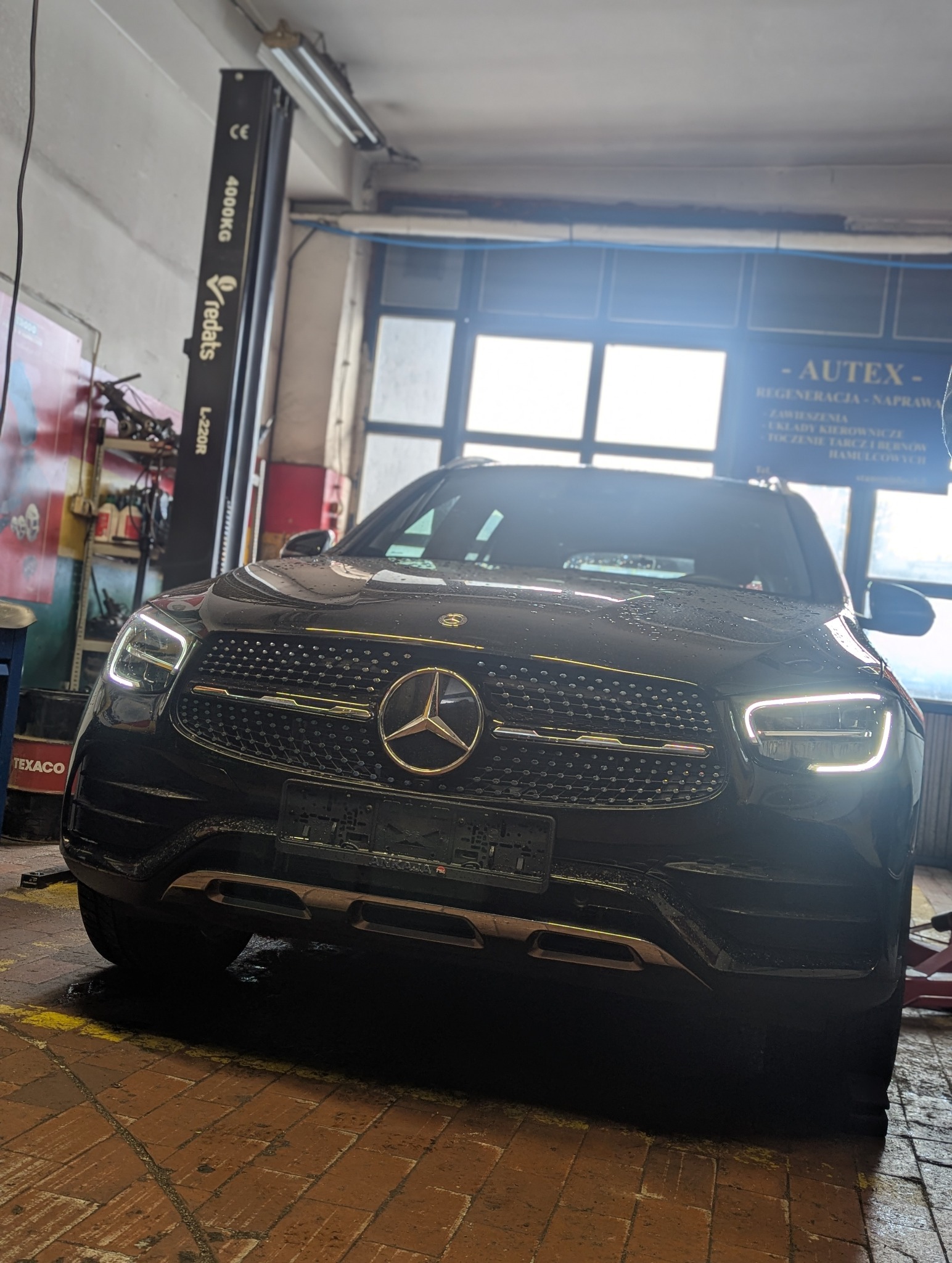 Czarny Mercedes-Benz GLC SUV z włączonymi światłami LED, stojący w warsztacie samochodowym na tle podnośnika i tablicy informacyjnej AUTEX.
