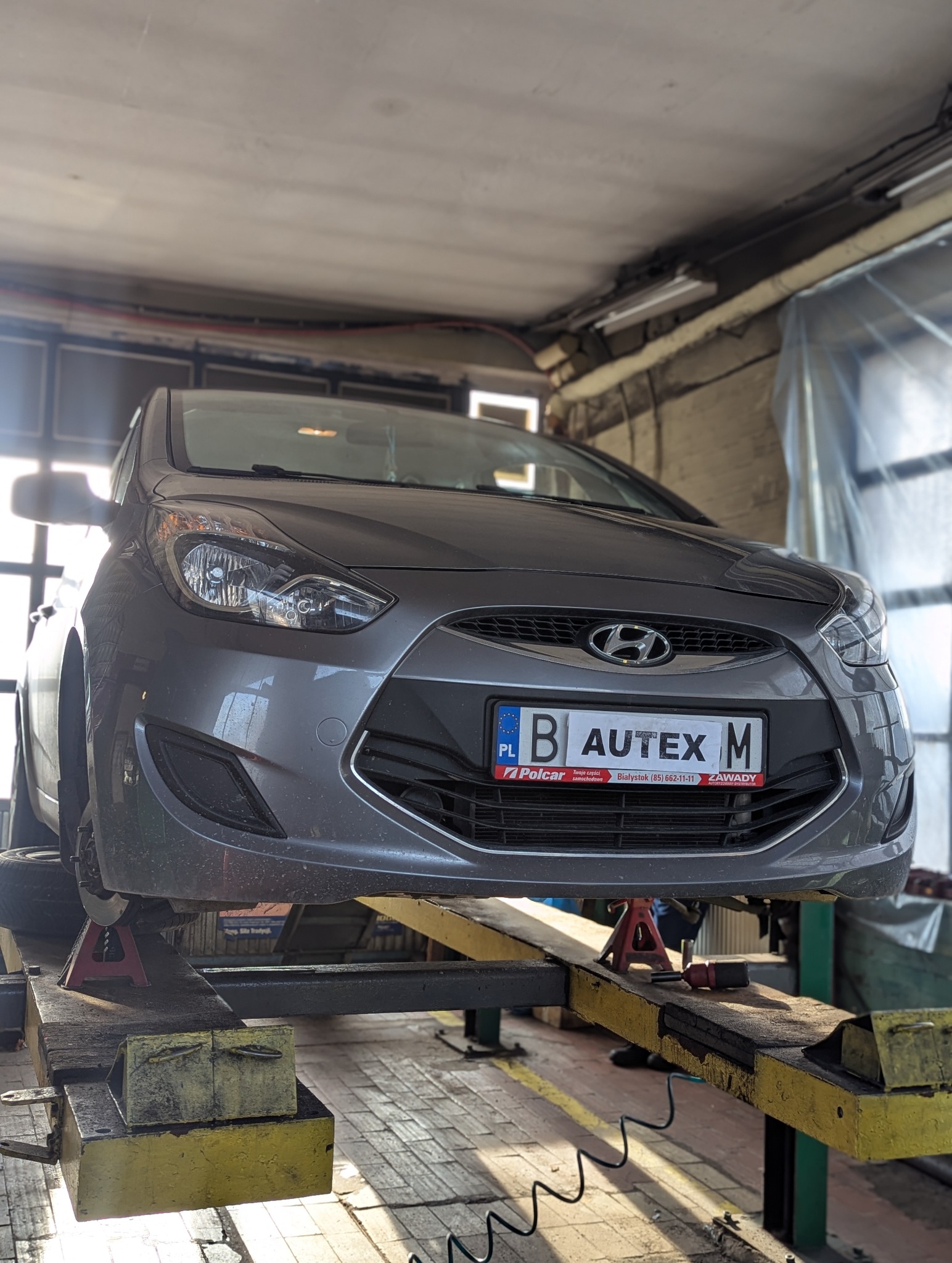 Szary samochód Hyundai na podnośniku warsztatowym, widoczne czerwone podpórki pod wahaczami, tablica rejestracyjna z Białegostoku.