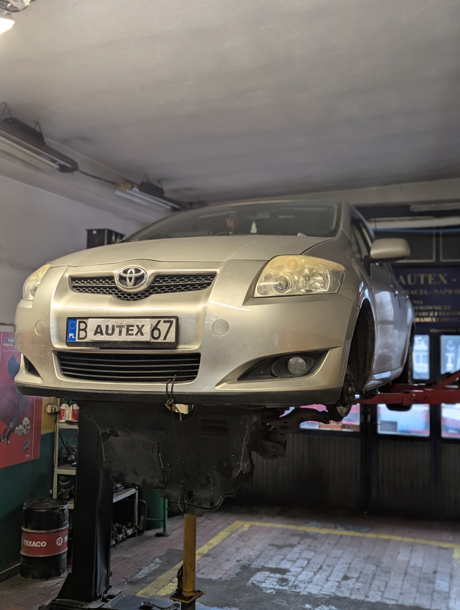 Srebrna Toyota Auris na podnośniku samochodowym w warsztacie, widoczna tablica rejestracyjna z napisem AUTEX 67, brak jednego koła, w tle ściana z reklamą warsztatu i beczka Texaco.