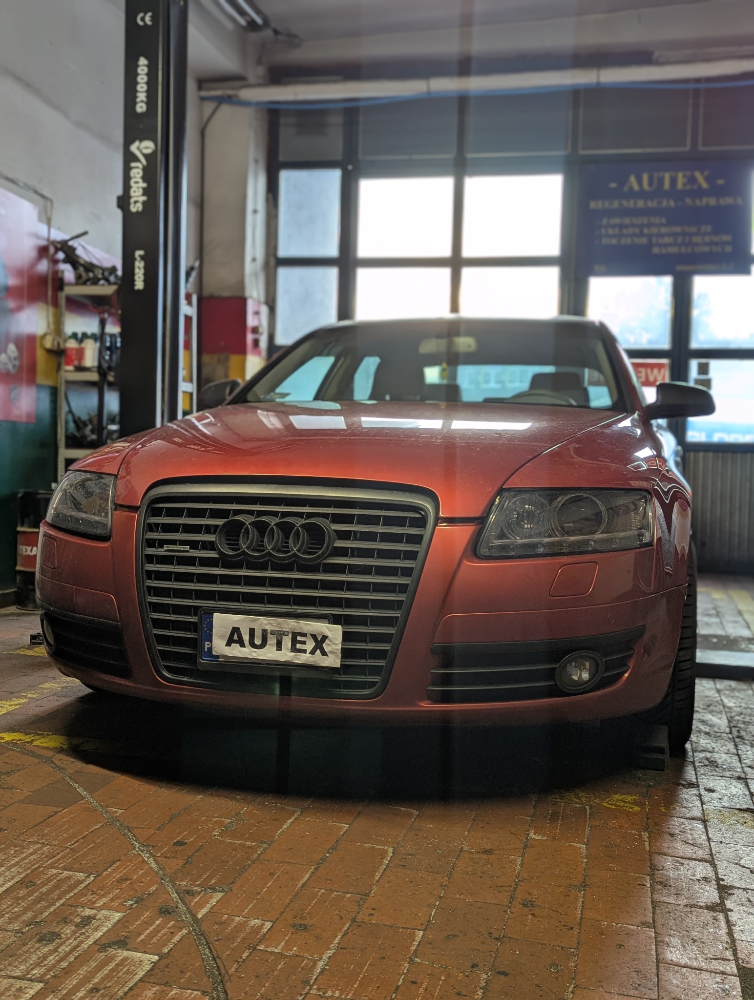 Czerwone Audi A6 na ceglanej podłodze warsztatu samochodowego, widoczny podnośnik kolumnowy i szyld z nazwą warsztatu 'Autex'.