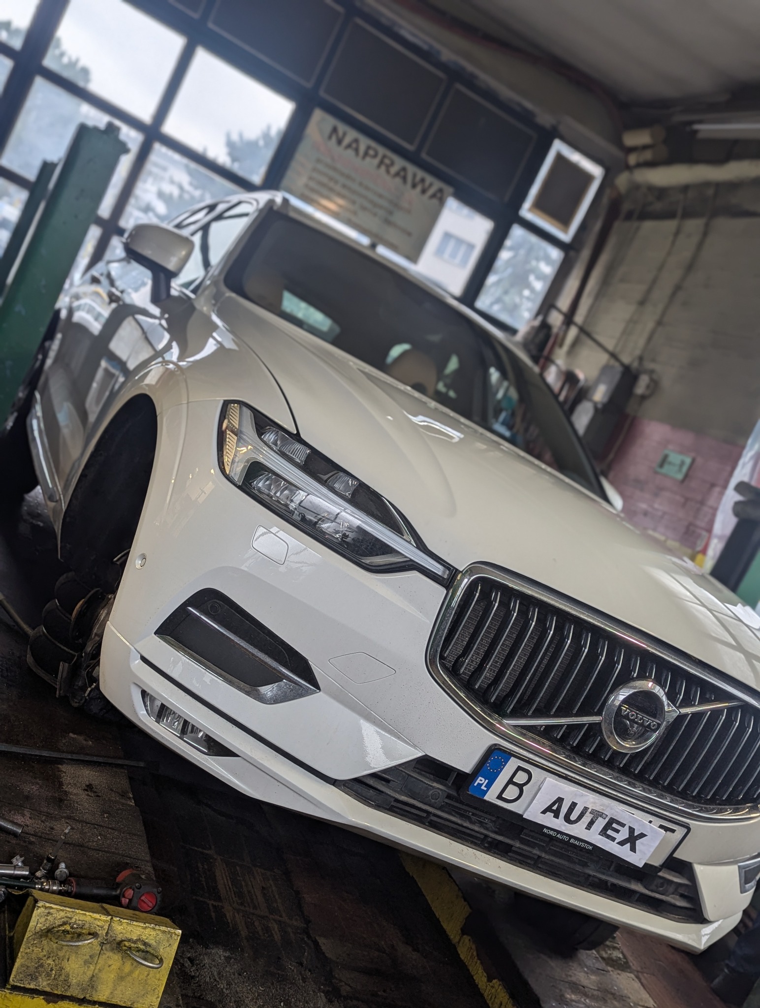 Białe auto marki Volvo na podnośniku w warsztacie, brak przedniego koła, widoczny numer rejestracyjny z naklejką AUTEX