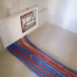 Rozdzielacz ogrzewania podłogowego w białej szafce z wystającymi rurami w kolorach czerwonym i niebieskim, ułożonymi równolegle na surowej betonowej posadzce.