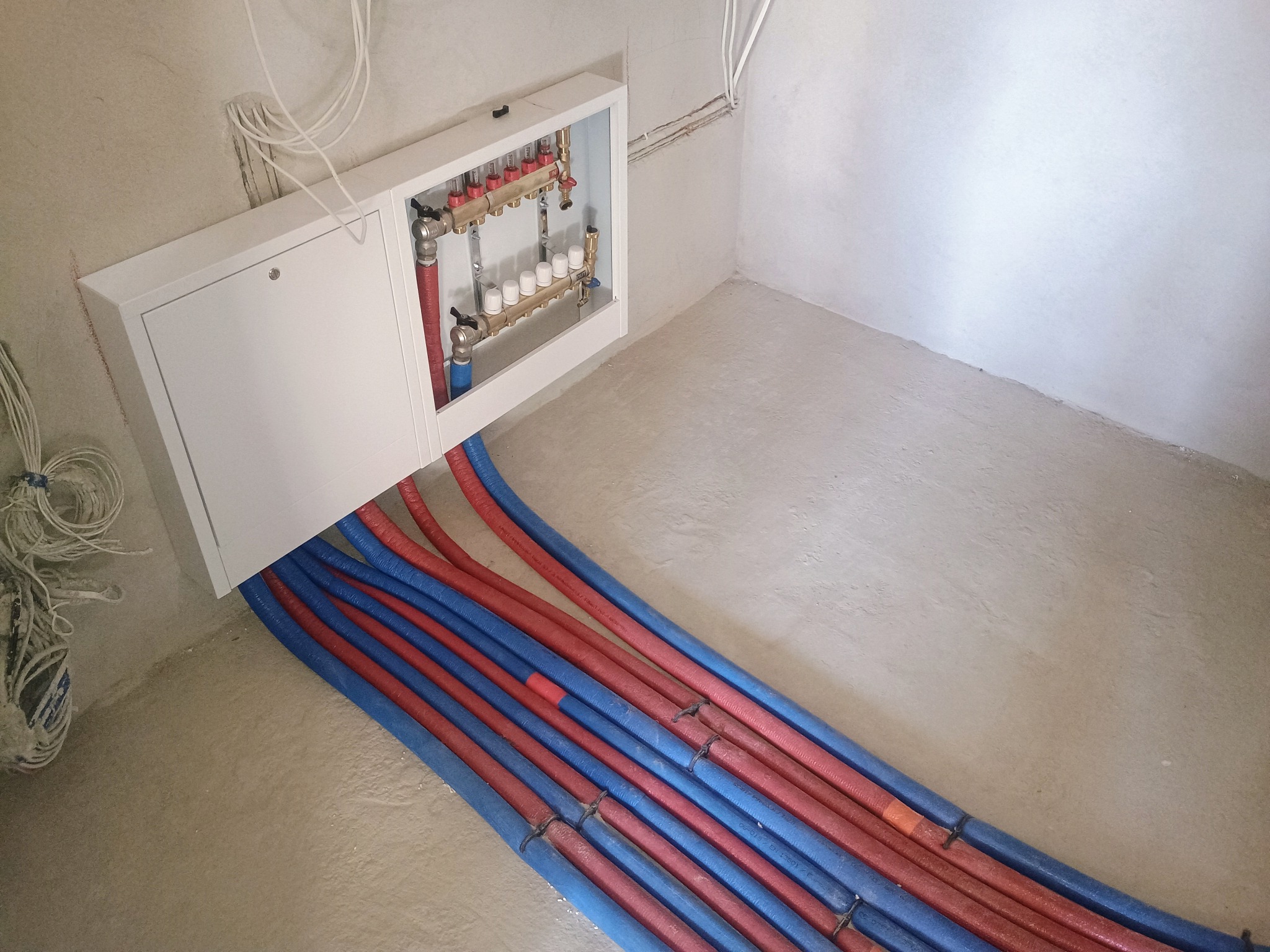 Rozdzielacz ogrzewania podłogowego w białej szafce z wystającymi rurami w kolorach czerwonym i niebieskim, ułożonymi równolegle na surowej betonowej posadzce.