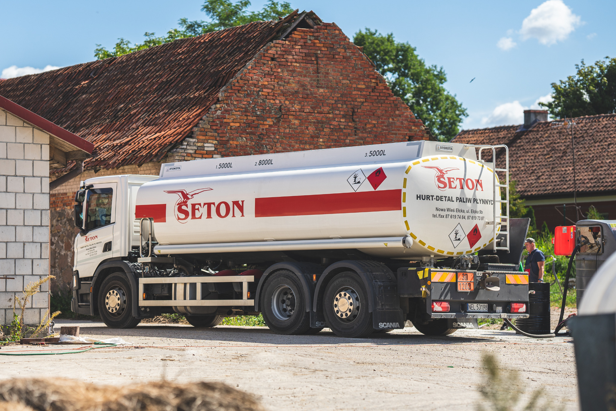 Cysterna z logo firmy SETON do transportu paliw płynnych, zaparkowana na tle budynku z czerwonej cegły.