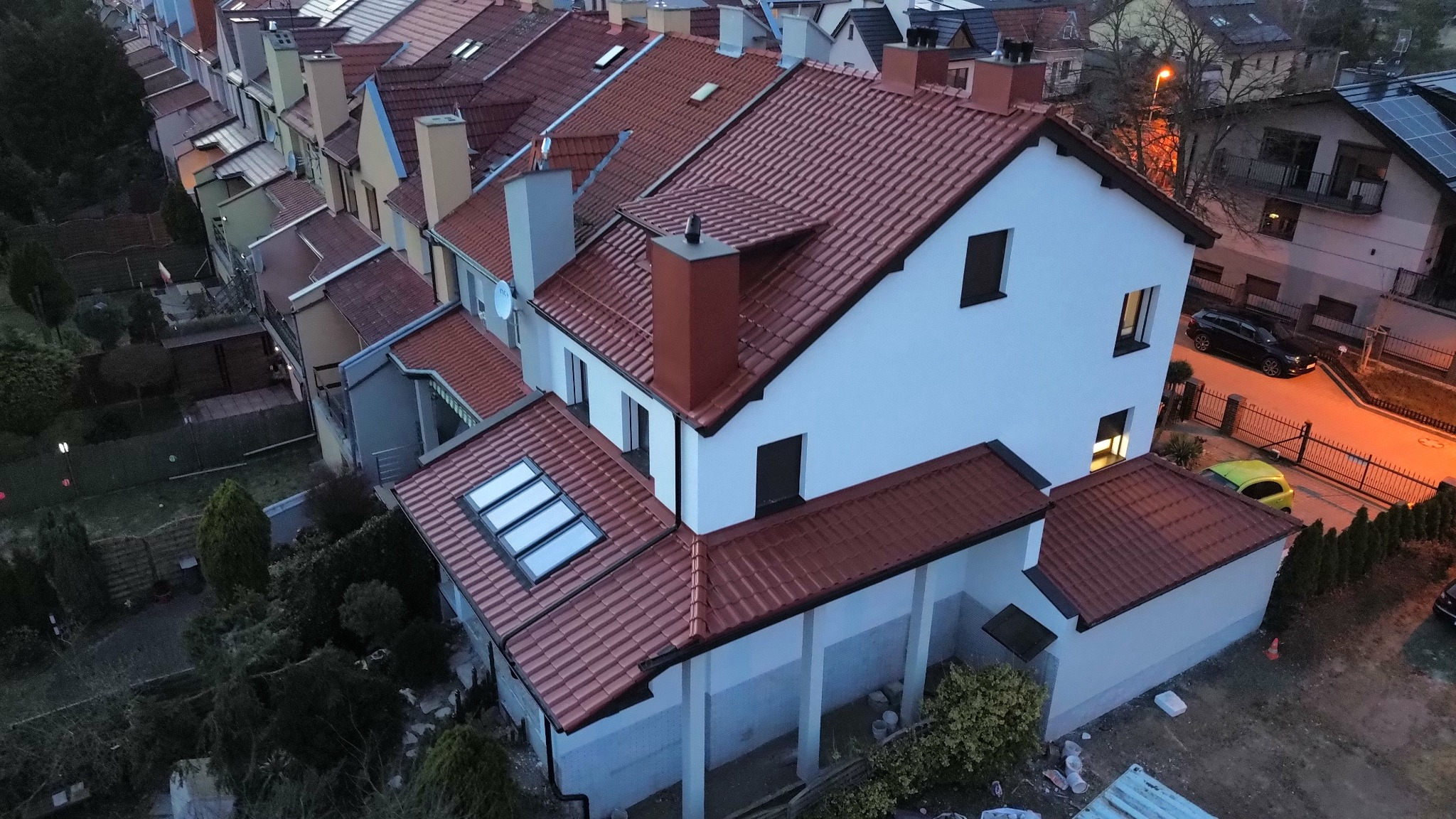 Widok z góry na szereg domów z nową, czerwoną dachówką ceramiczną, komin pomalowany w kolorze dachu, okna dachowe, biała elewacja budynku, widoczna zabudowa sąsiednia.