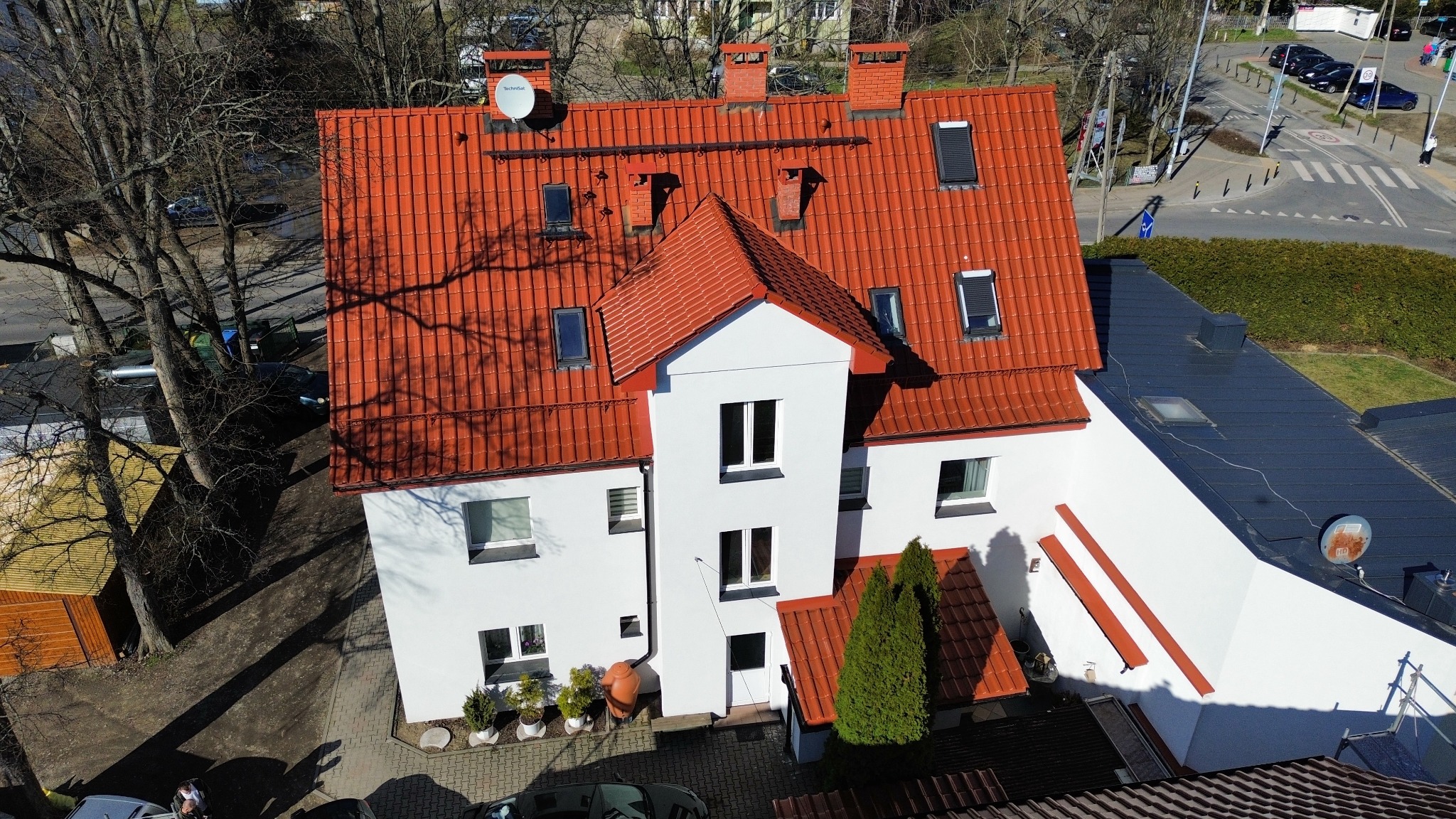 Widok z góry na dom z czerwoną dachówką, kominami i anteną satelitarną, otoczony zielenią i drzewami w słoneczny dzień.