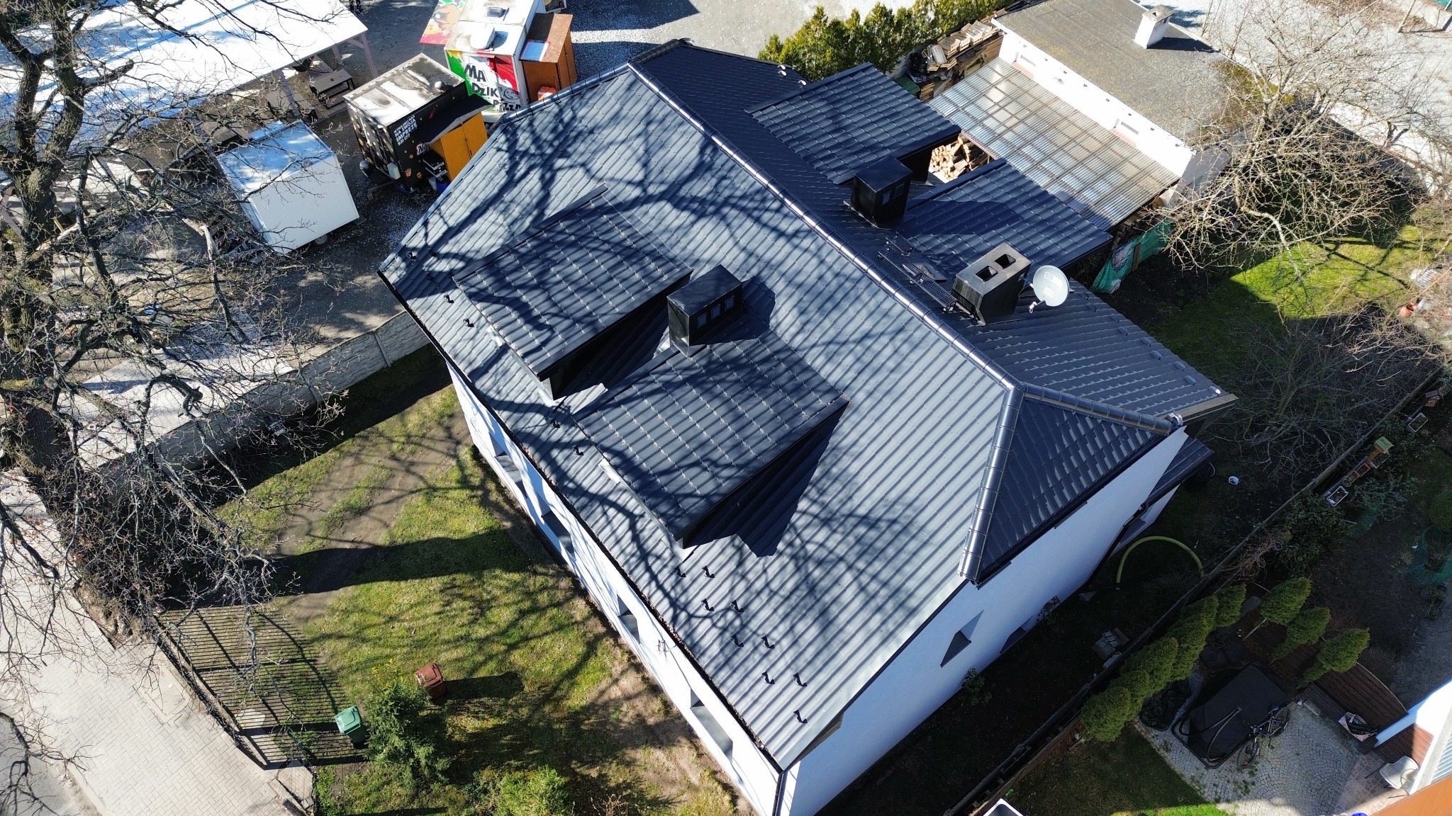 Widok z góry na dom z ciemnoszarą dachówką ceramiczną, z trzema oknami dachowymi i kominem, cień drzewa na dachu, biała elewacja, zielony trawnik, w tle zabudowania gospodarcze i drzewa.