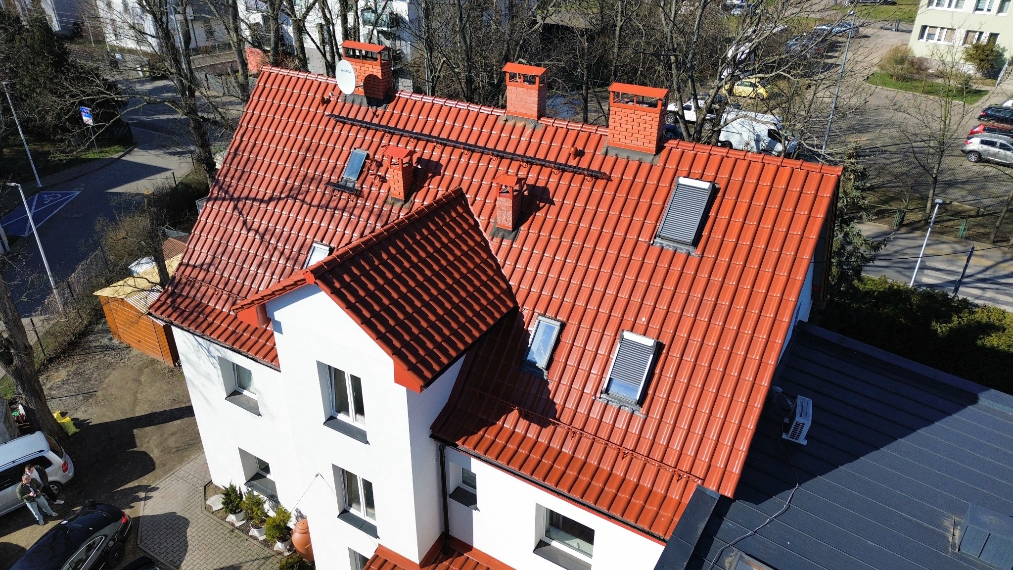 Widok z góry na odnowiony dach z czerwonej dachówki ceramicznej z lukarnami i kominami, biała elewacja budynku, otoczenie miejskie.