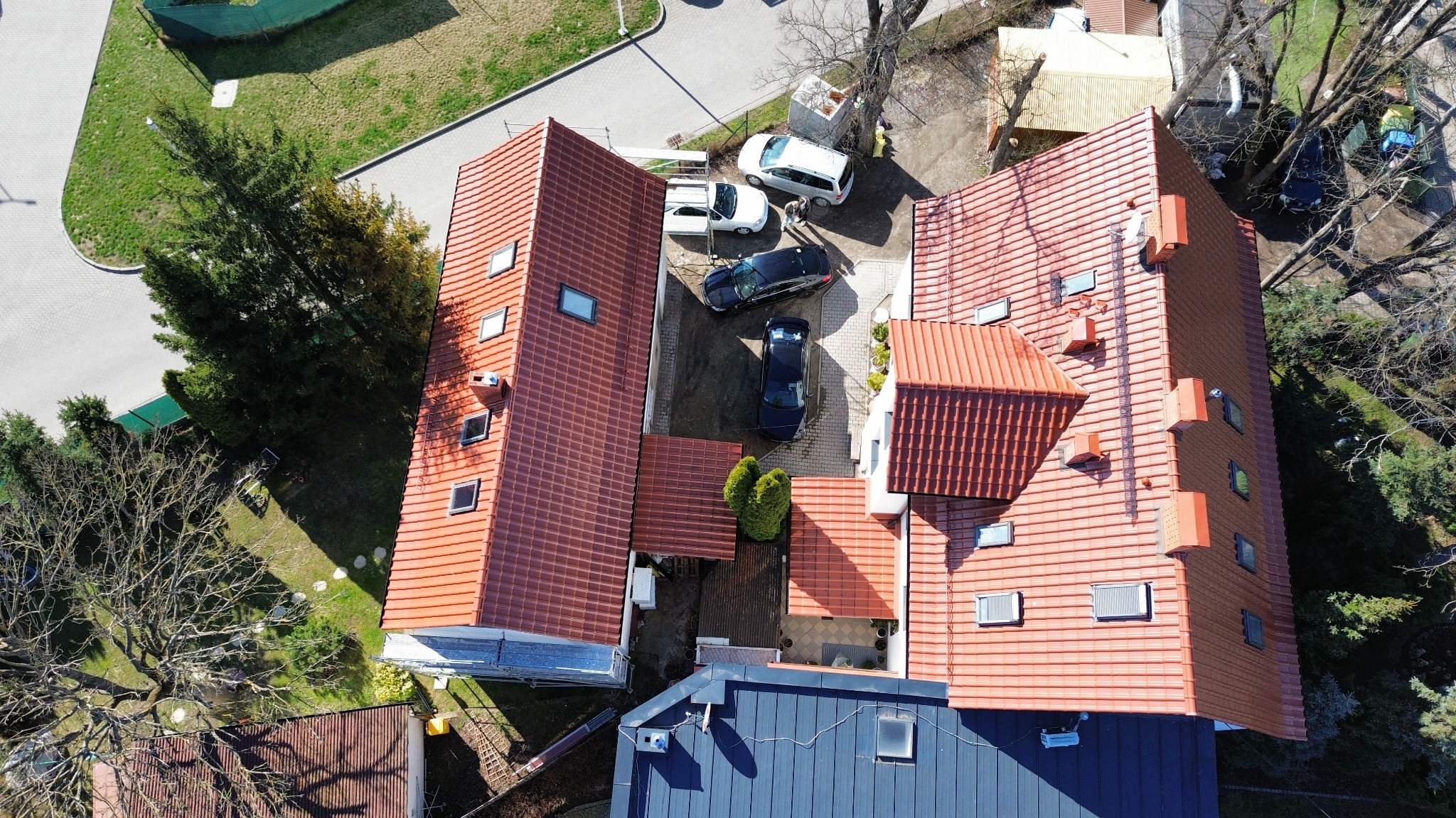 Widok z góry na dwa budynki z dachami pokrytymi czerwoną dachówką, pomiędzy nimi zaparkowane samochody, otoczenie zielone z drzewami.