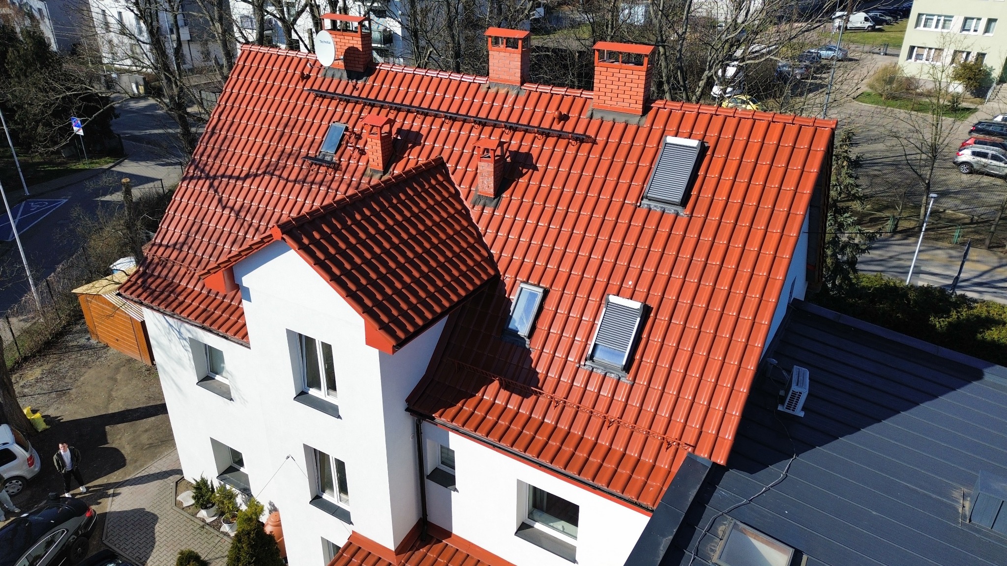 Widok z góry na dom z nową, czerwoną dachówką ceramiczną, trzema kominami z cegły, oknami dachowymi z roletami i anteną satelitarną. Widoczna część sąsiedniego dachu pokrytego blachą trapezową...