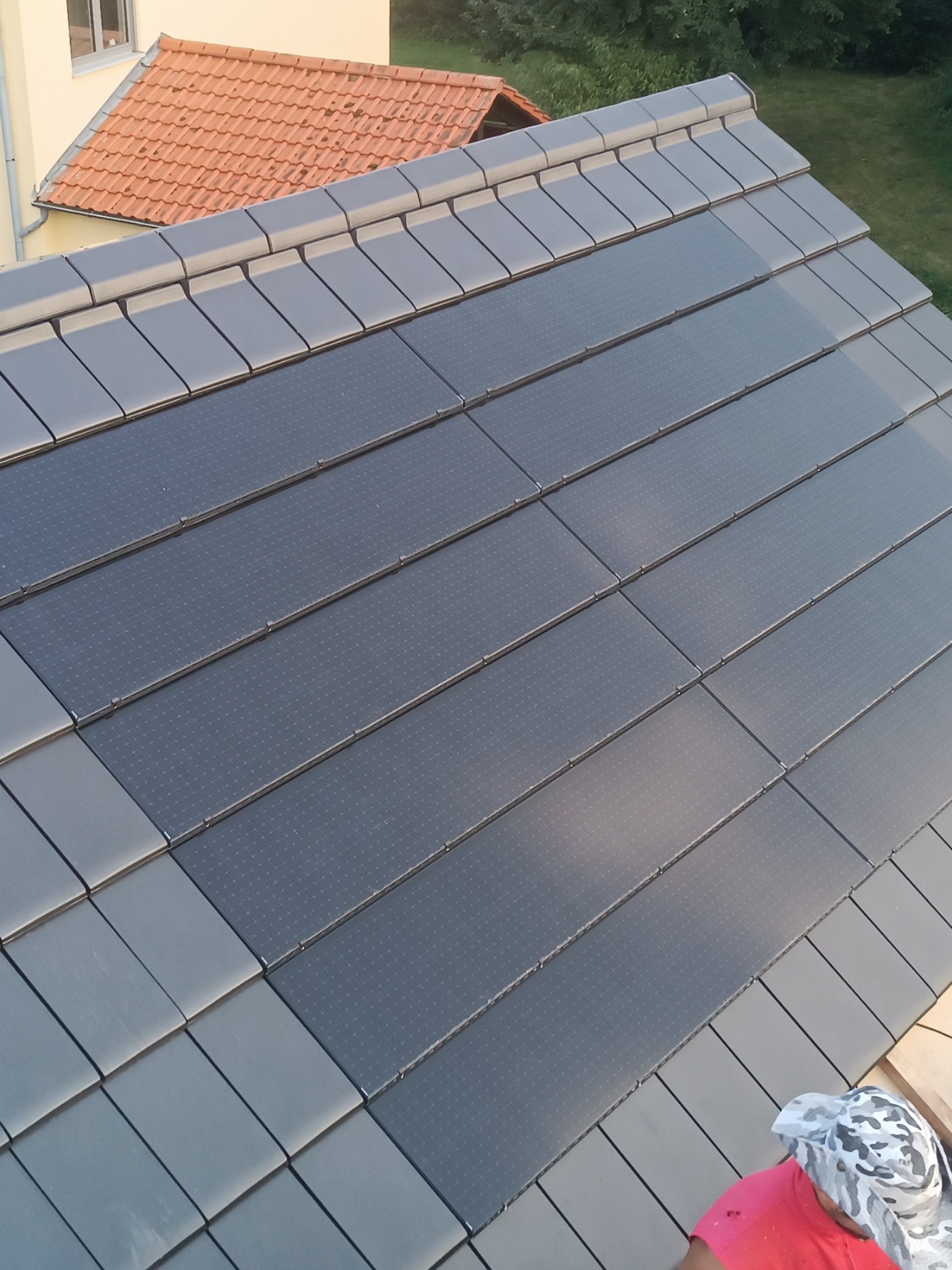 Ukośny widok na fragment dachu pokrytego szarymi dachówkami z wbudowanymi panelami solarnymi; w tle widoczny fragment pomarańczowego dachu innego budynku i zieleń.