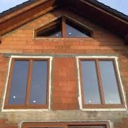 White Energy Przemysław Lis Okna Pro + - Surowy stan elewacji domu z widocznymi dwoma nowo zamontowanymi oknami z brązowymi ramami i trójkątnym oknem na poddaszu, widoczne ślady zaprawy murarskiej.