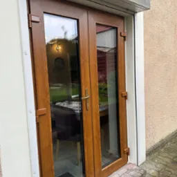 Zewnętrzne dwuskrzydłowe drzwi balkonowe z brązowego tworzywa, z widoczną częściowo zwiniętą roletą zewnętrzną nad nimi, zamontowane w otynkowanym budynku.