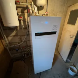 Biała pompa ciepła Panasonic Aquarea Smart Cloud zamontowana w kotłowni w Poznaniu, widoczne liczne rury i przyłącza, obok stoi biały bojler Tesy.