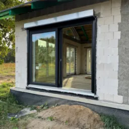 Czarne okno przesuwne z roletą zewnętrzną w surowym stanie budynku z betonu komórkowego. Widoczne wnętrze i fragment trawnika.