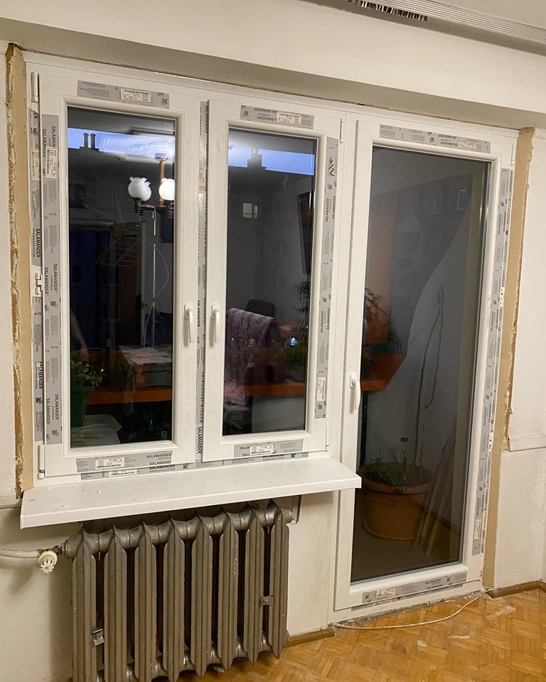 Montaż okna PCV Salamander, widoczne taśmy zabezpieczające na ramie, stary grzejnik pod oknem i fragment parkietu.