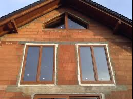 Surowy stan elewacji domu z widocznymi dwoma nowo zamontowanymi oknami z brązowymi ramami i trójkątnym oknem na poddaszu, widoczne ślady zaprawy murarskiej.