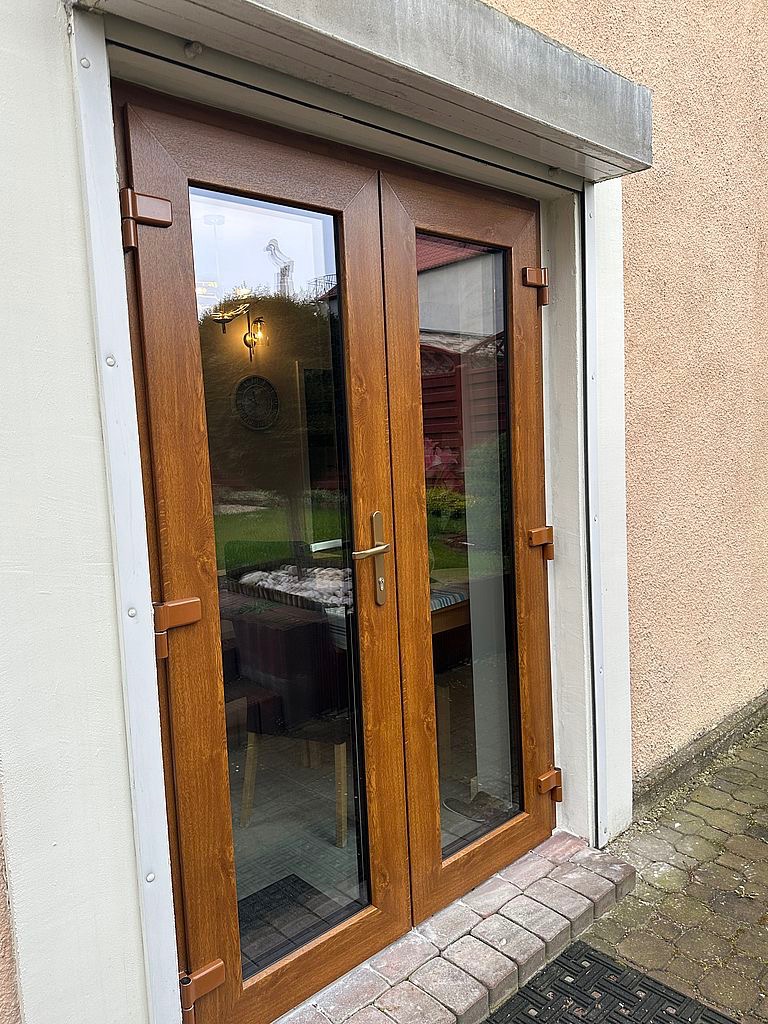 Zewnętrzne dwuskrzydłowe drzwi balkonowe z brązowego tworzywa, z widoczną częściowo zwiniętą roletą zewnętrzną nad nimi, zamontowane w otynkowanym budynku.