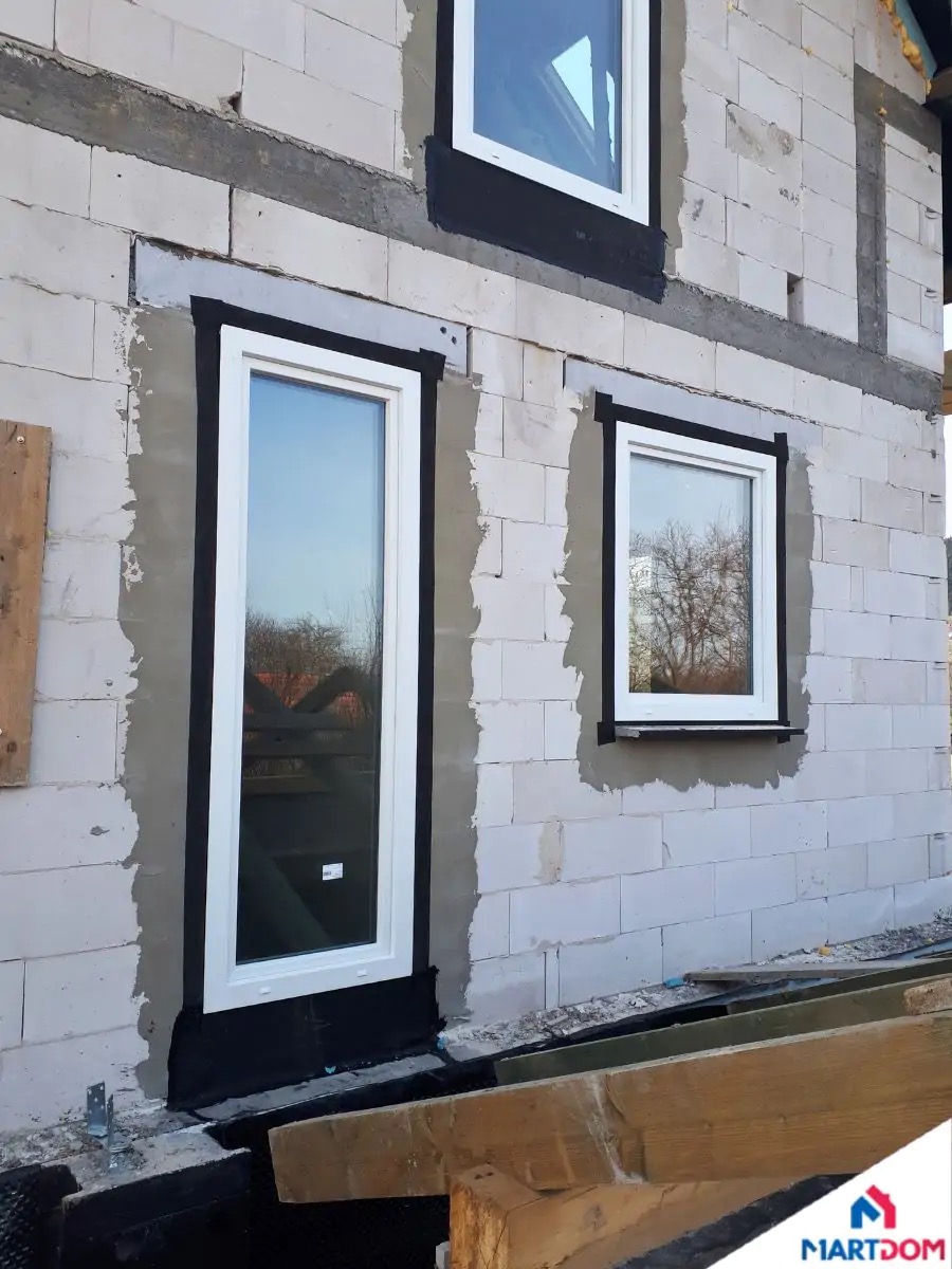 Wstawione okna i drzwi balkonowe w surowej ścianie z betonu komórkowego, widoczne czarne taśmy uszczelniające i świeże obramowanie cementowe, drewniane stemple pod oknem balkonowym.