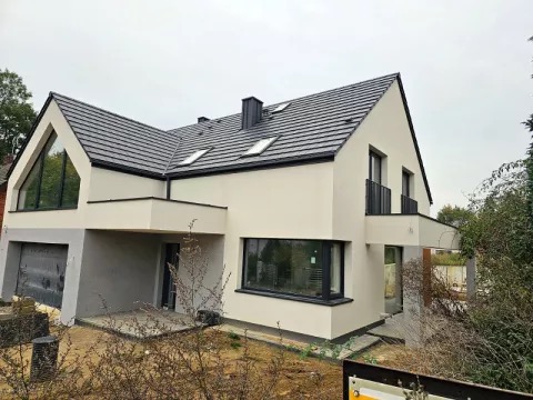 Nowoczesny dom jednorodzinny z dużym trójkątnym oknem, balkonem z czarną balustradą i szarą dachówką, widoczny w pochmurny dzień.