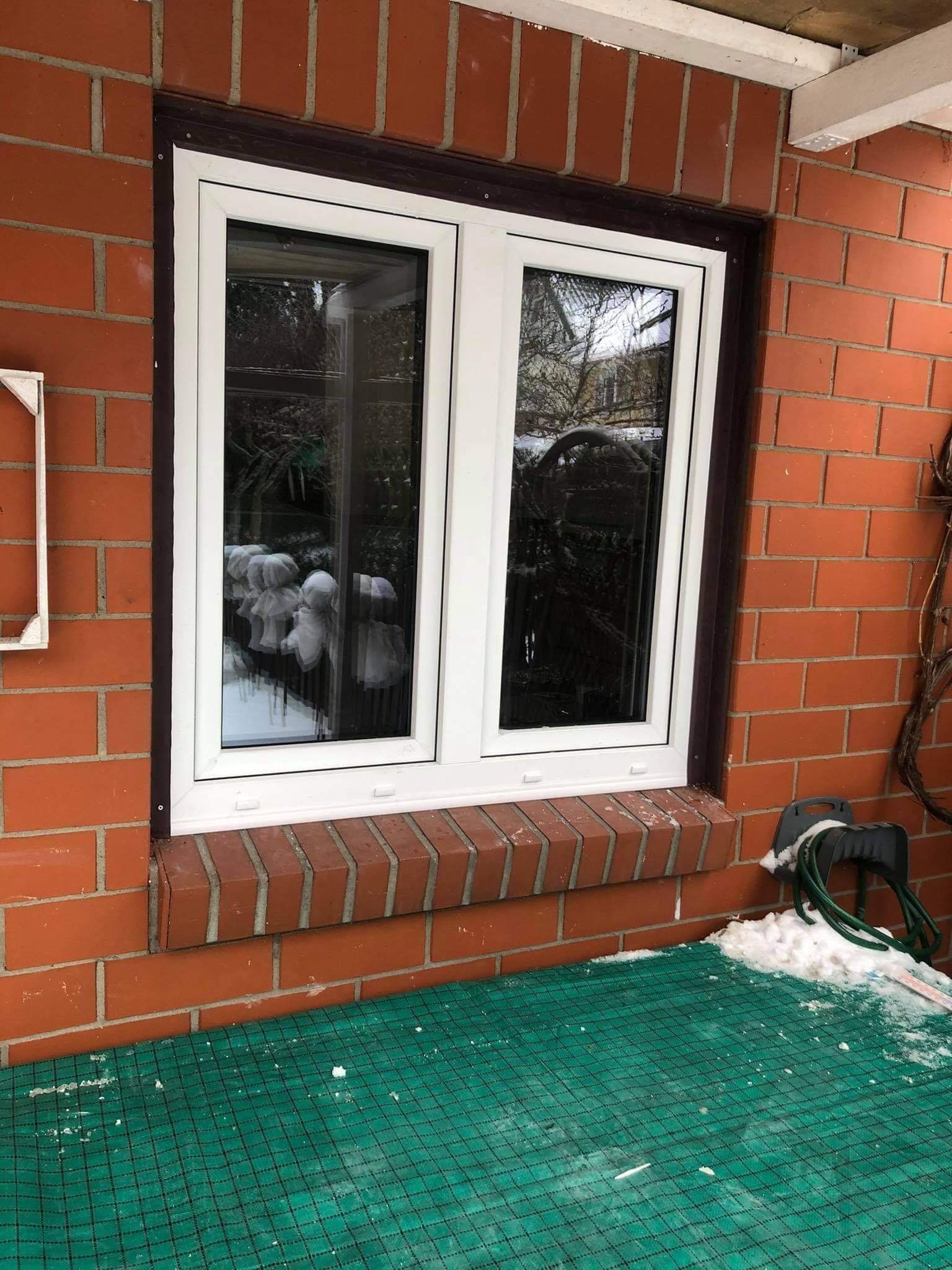 Białe, dwuskrzydłowe okno wstawione w ceglaną ścianę, widok z zewnątrz, na parapecie ceglanym leży śnieg, a w oknie odbijają się ozdoby ogrodowe.