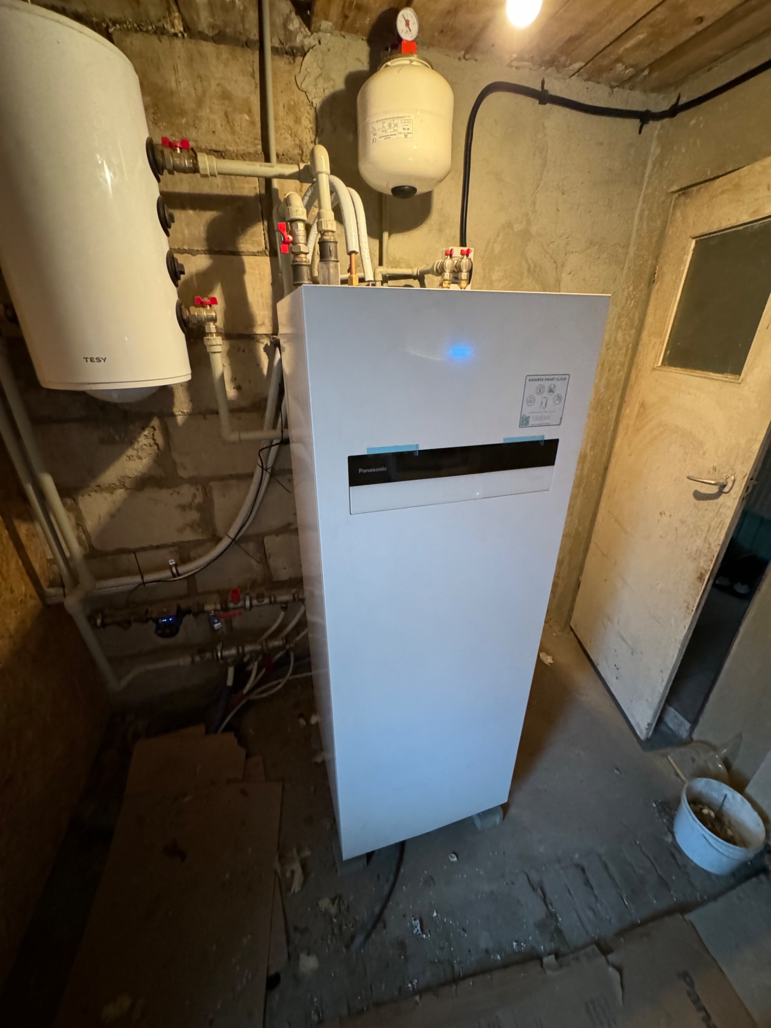 Biała pompa ciepła Panasonic Aquarea Smart Cloud zamontowana w kotłowni w Poznaniu, widoczne liczne rury i przyłącza, obok stoi biały bojler Tesy.