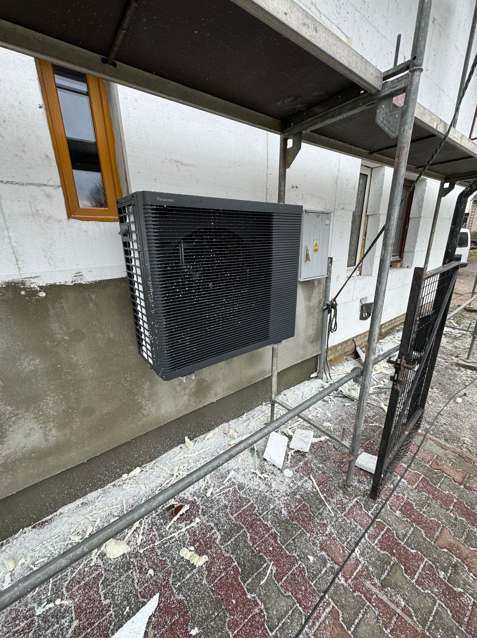 Czarna jednostka zewnętrzna pompy ciepła Panasonic zamontowana na ocieplonym budynku w trakcie prac wykończeniowych, widoczne rusztowanie i resztki materiałów budowlanych.