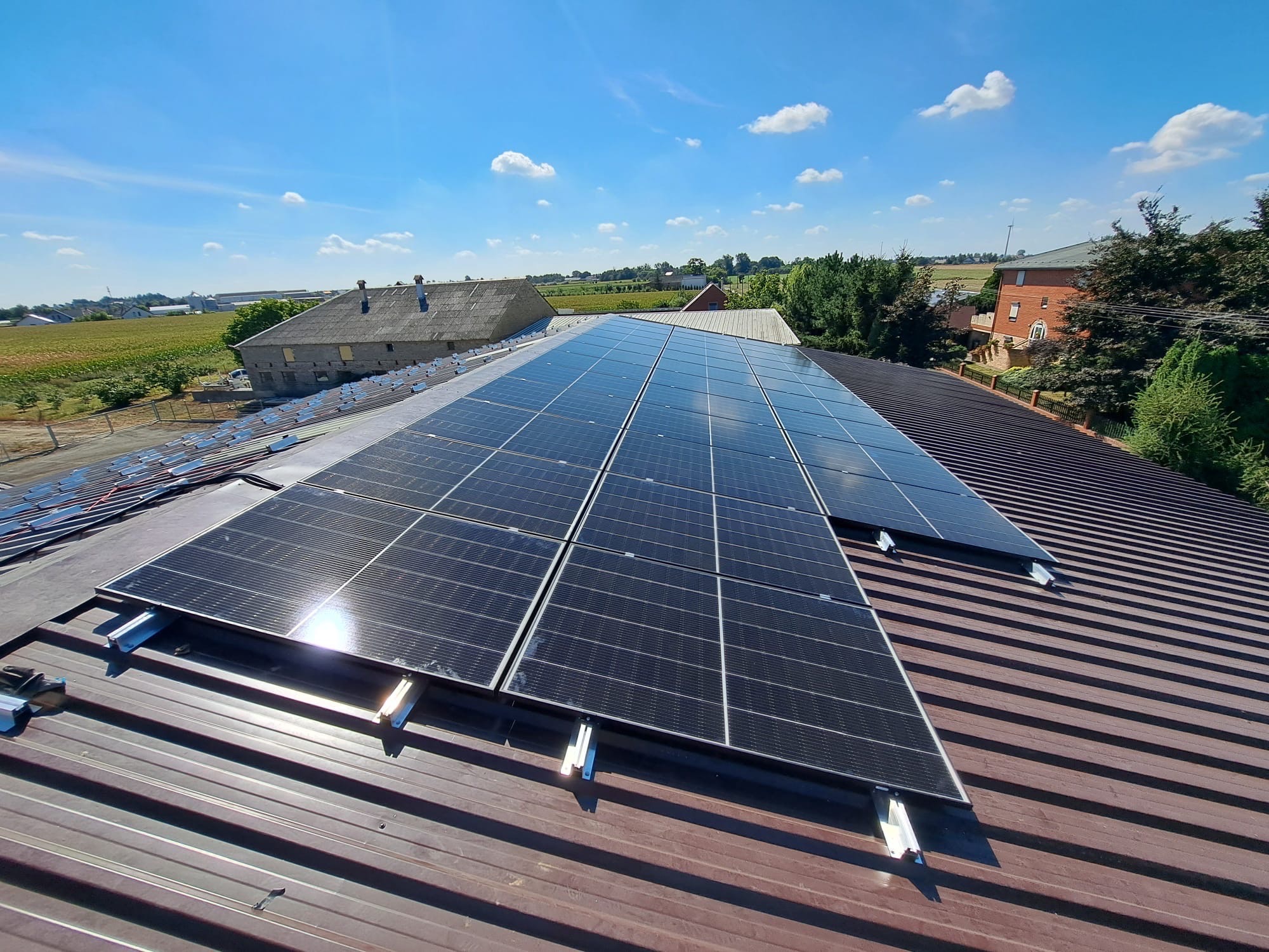 Instalacja paneli słonecznych na brązowym dachu blaszanym, z widokiem na okoliczne budynki i błękitne niebo z chmurami w tle. Widoczne elementy montażowe.