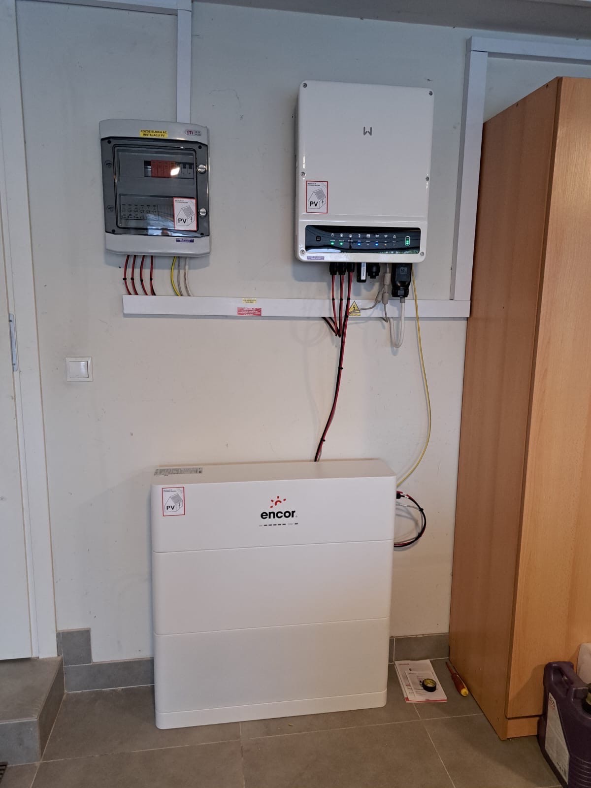 Kompletna instalacja fotowoltaiczna wewnątrz pomieszczenia: rozdzielnica, inwerter i akumulator energii Encor, zamontowane na ścianie. Widoczne przewody i elementy montażowe.
