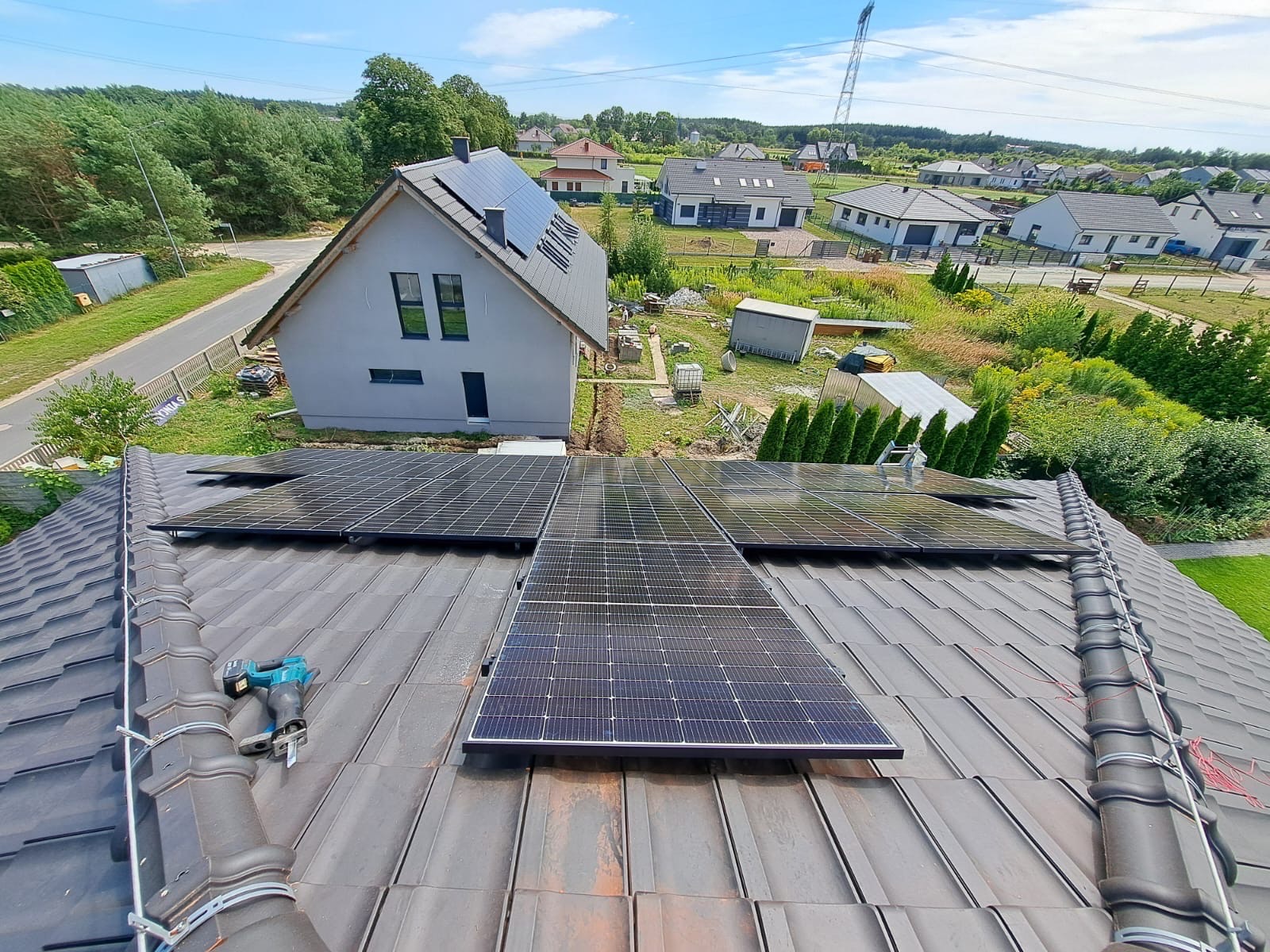 Montaż paneli fotowoltaicznych na dachu domu jednorodzinnego z użyciem narzędzi. Widok z góry, w tle zabudowa mieszkalna i zieleń. Dachówka ceramiczna.