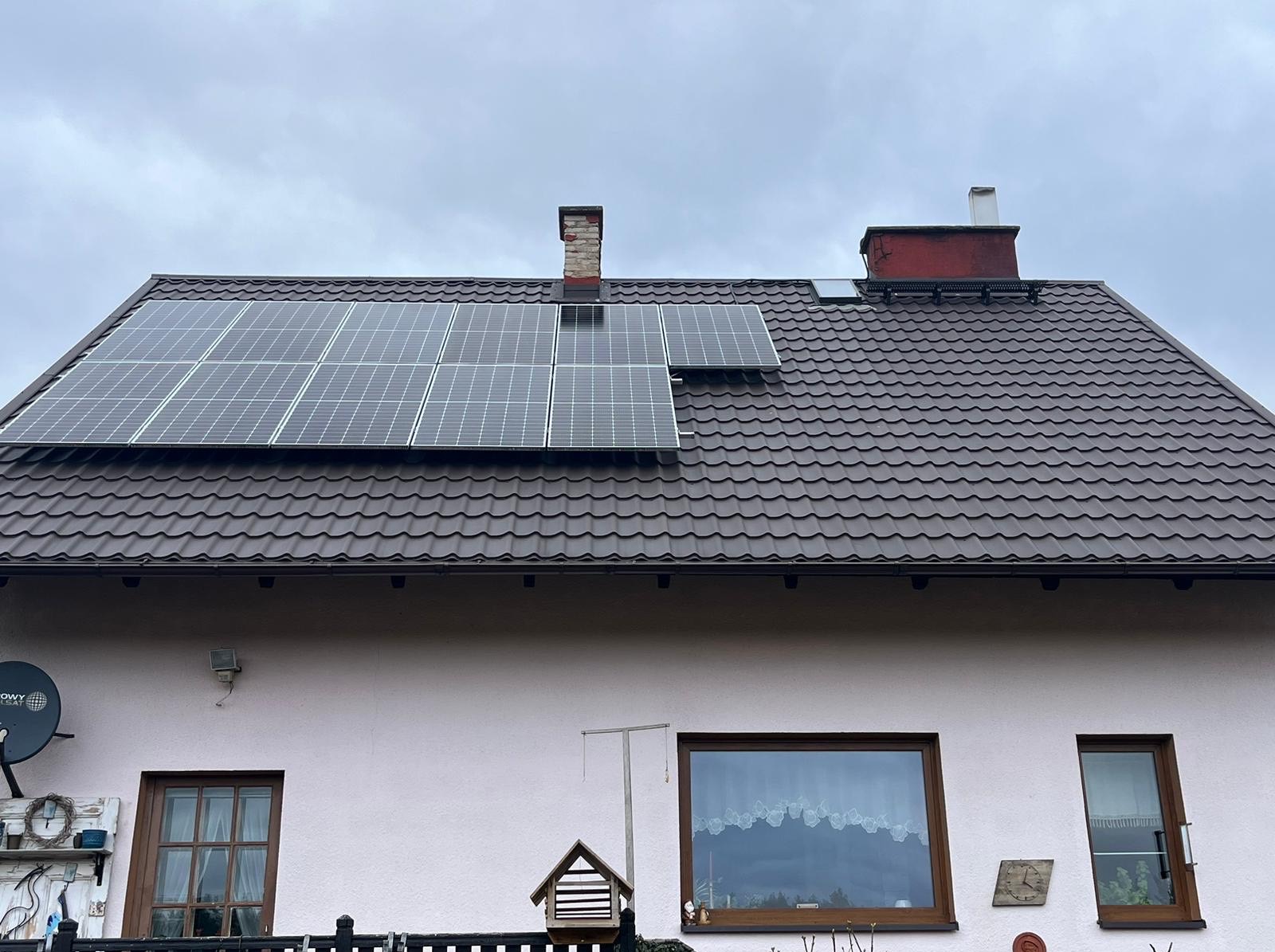 Brązowy dach z panelami słonecznymi na jasnym domu jednorodzinnym w pochmurny dzień. Widoczne okna z dekoracjami i antena satelitarna.