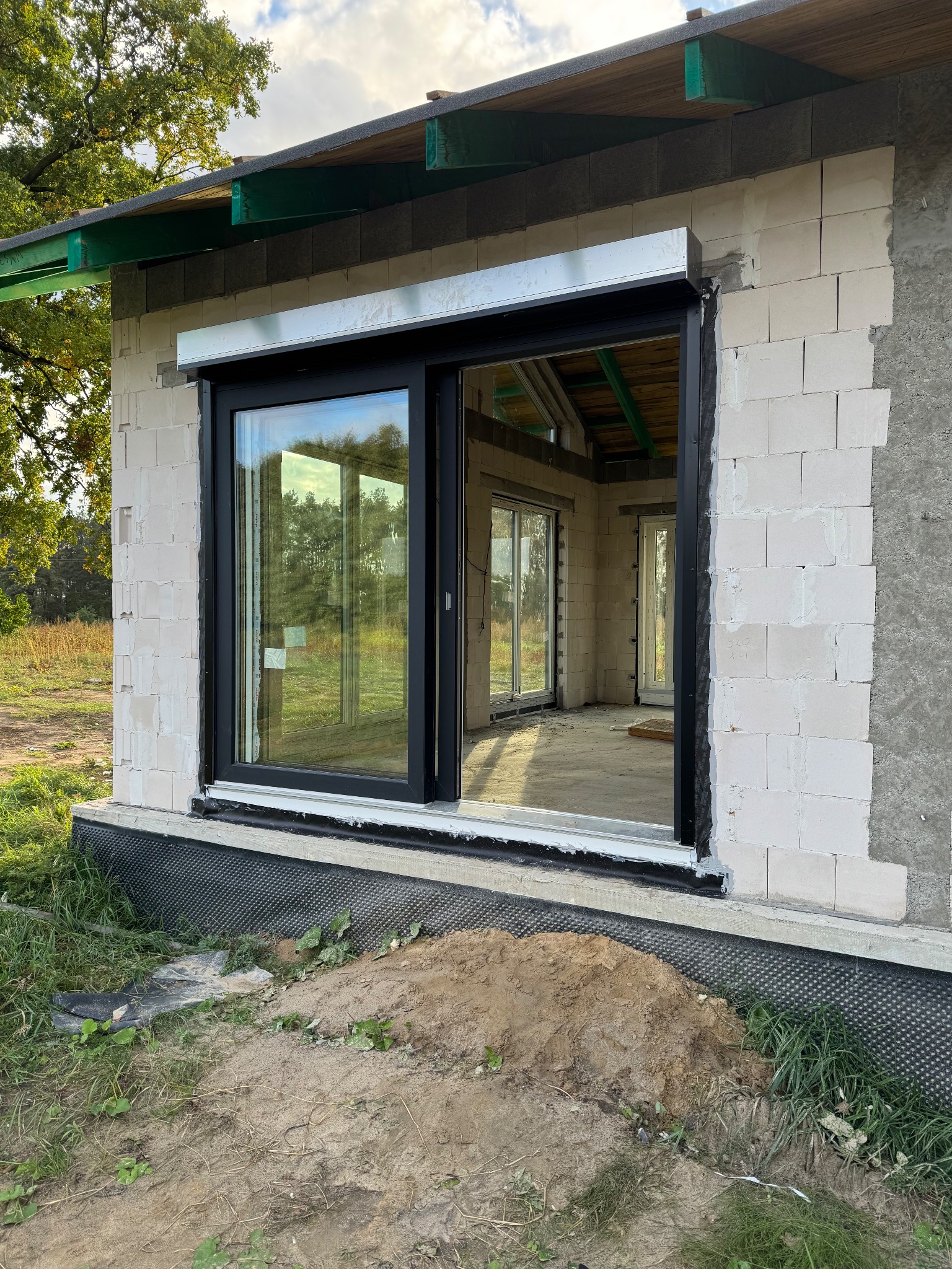 Czarne okno przesuwne z roletą zewnętrzną w surowym stanie budynku z betonu komórkowego. Widoczne wnętrze i fragment trawnika.
