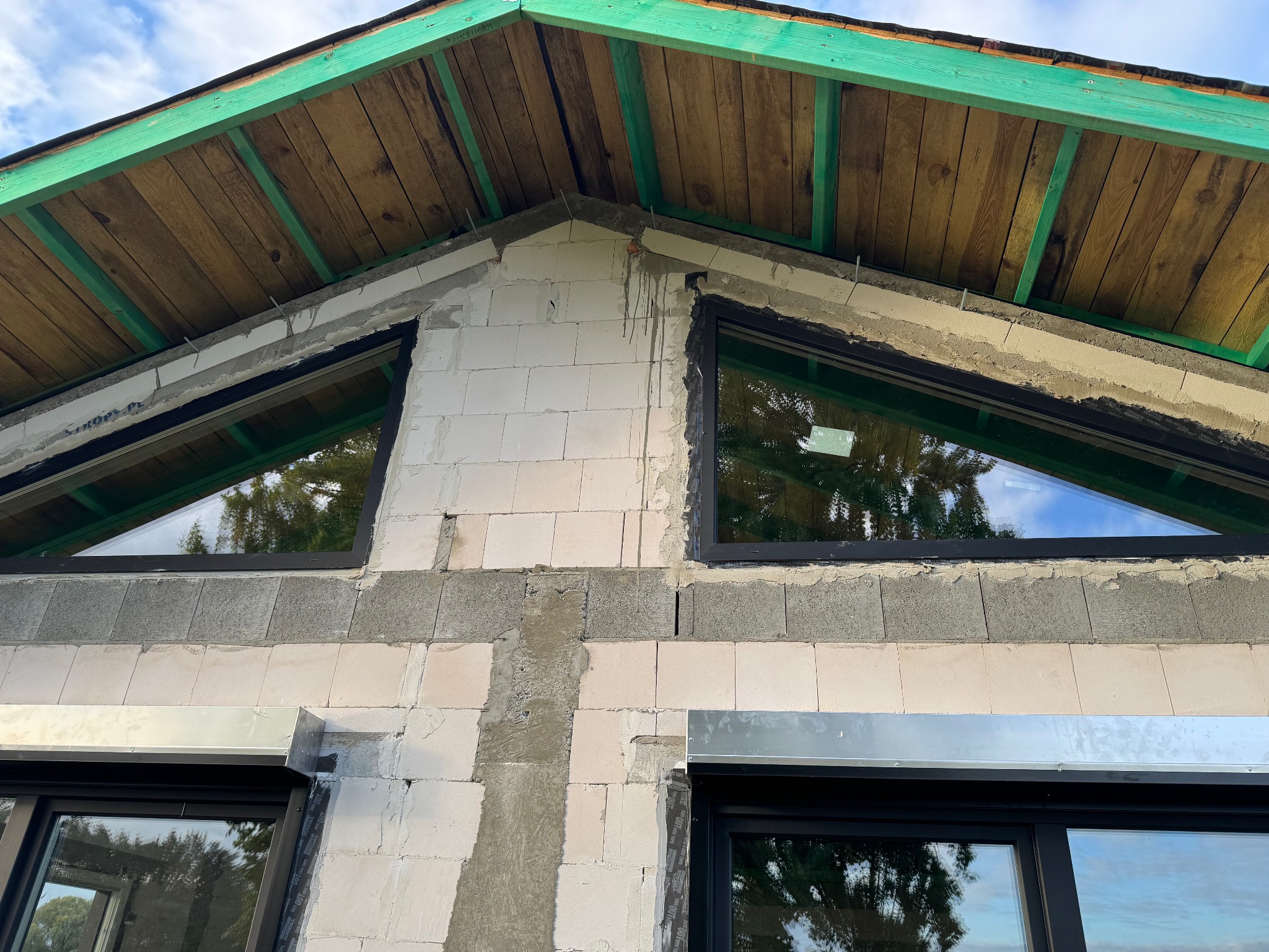 Surowy stan budynku z zamontowanymi czarnymi oknami i roletą zewnętrzną na parterze, widok na fragment ściany szczytowej i drewnianą konstrukcję dachu.