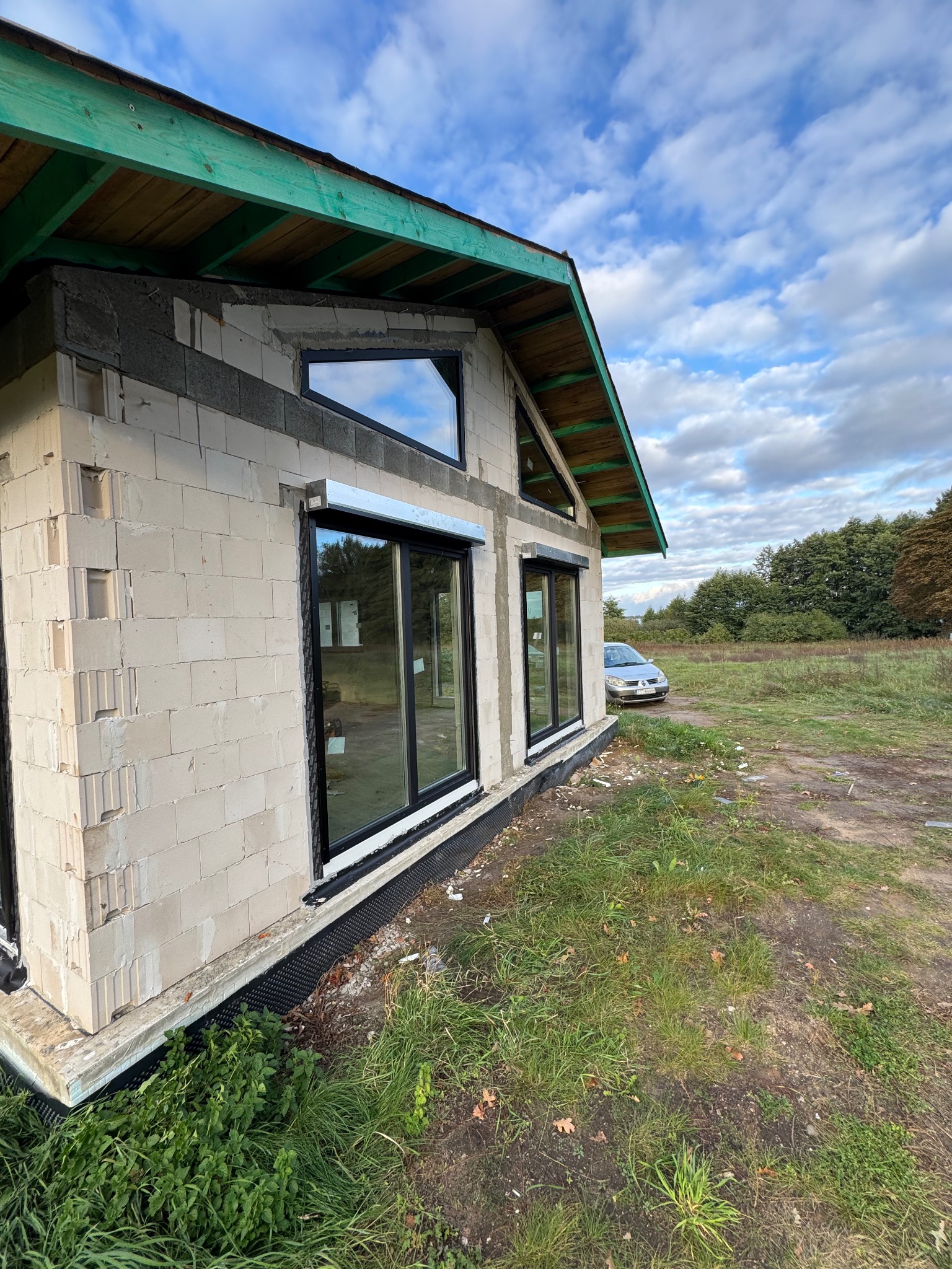 Surowy stan budynku z szarego betonu komórkowego z zamontowanymi oknami z czarnymi ramami i roletami zewnętrznymi. Widok z zewnątrz, w tle łąka i auto.