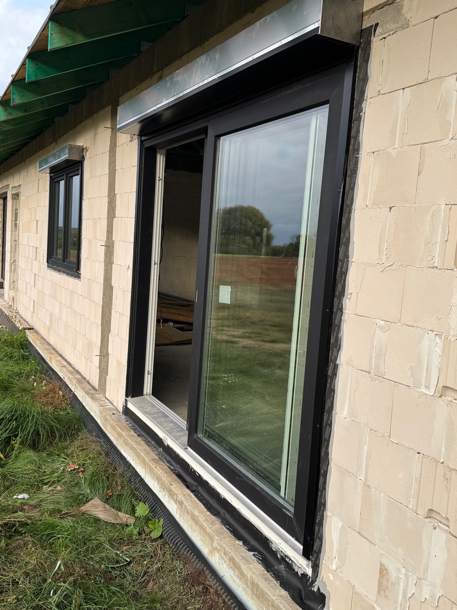 Czarne okno przesuwne z roletą zewnętrzną w surowym murze. Widoczne taśmy uszczelniające i fragment zielonego dachu. Niska perspektywa.