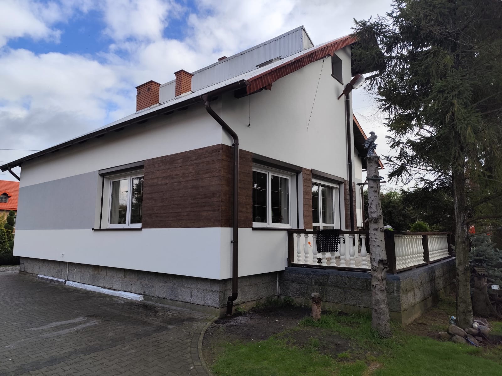 Elewacja domu z oknami i roletą, z brązowymi panelami imitującymi drewno. Widoczny fragment balkonu z białą balustradą i dekoracyjną figurką orła.