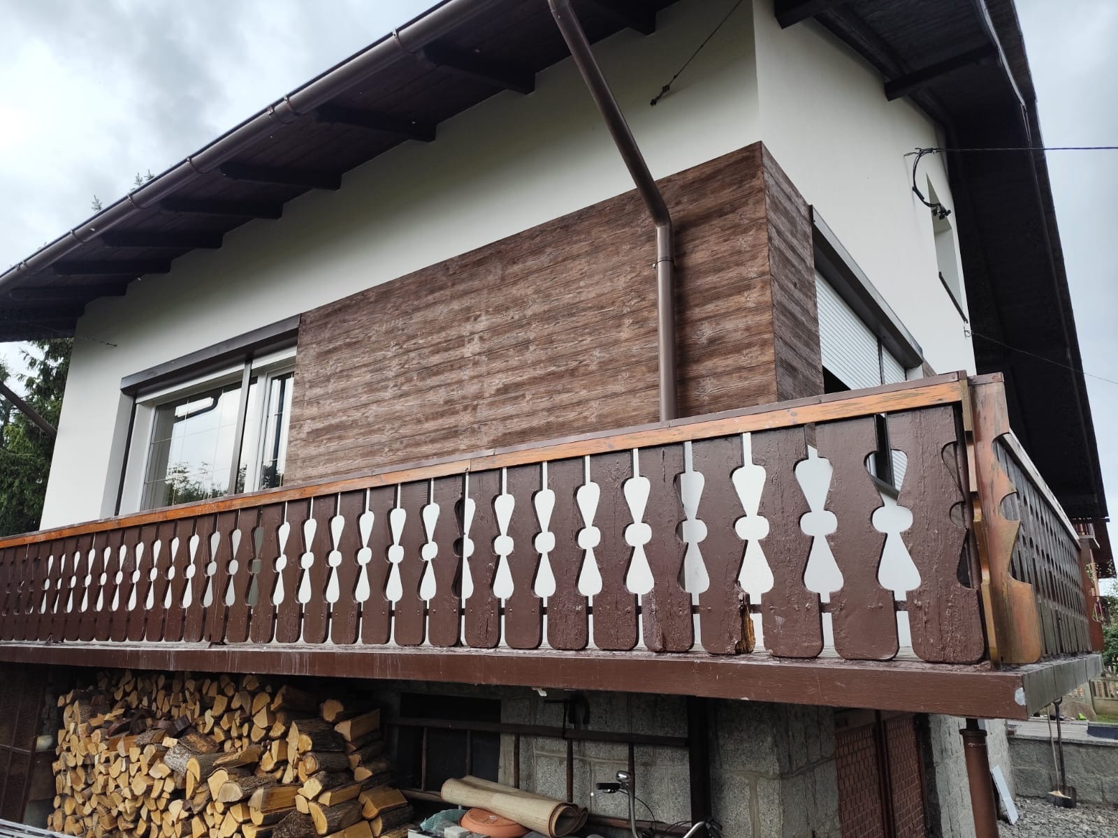 Balkon z drewnianą balustradą i fasadą imitującą drewno, uzupełniony o rolety okienne, tworzy przytulny wygląd domu z zapasem drewna pod spodem.