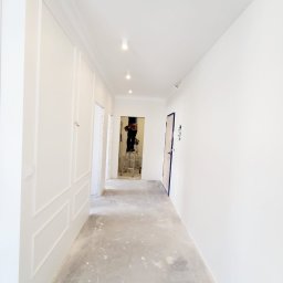 Presko - Korytarz w trakcie malowania na biało, widoczny pracownik na drabinie, świeżo pomalowane ściany z dekoracyjnymi panelami i sztukaterią, surowa podłoga zabezpieczona folią.