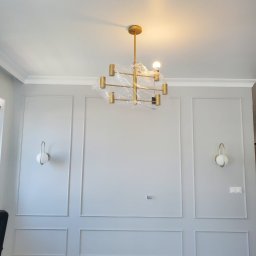 Presko - Szara ściana z dekoracyjnymi listwami, dwoma kinkietami z kulistymi kloszami i złotym, nowoczesnym żyrandolem w folii ochronnej, widoczne gniazdka elektryczne.