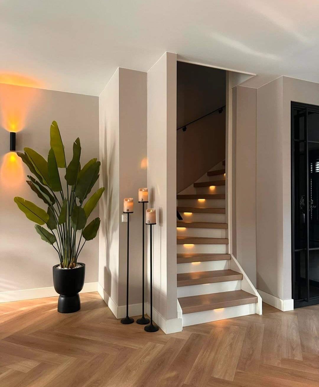 Jasny korytarz z parkietem w jodełkę, schodami z podświetleniem LED, wysoką rośliną w czarnej donicy i trzema świecami na czarnych stojakach